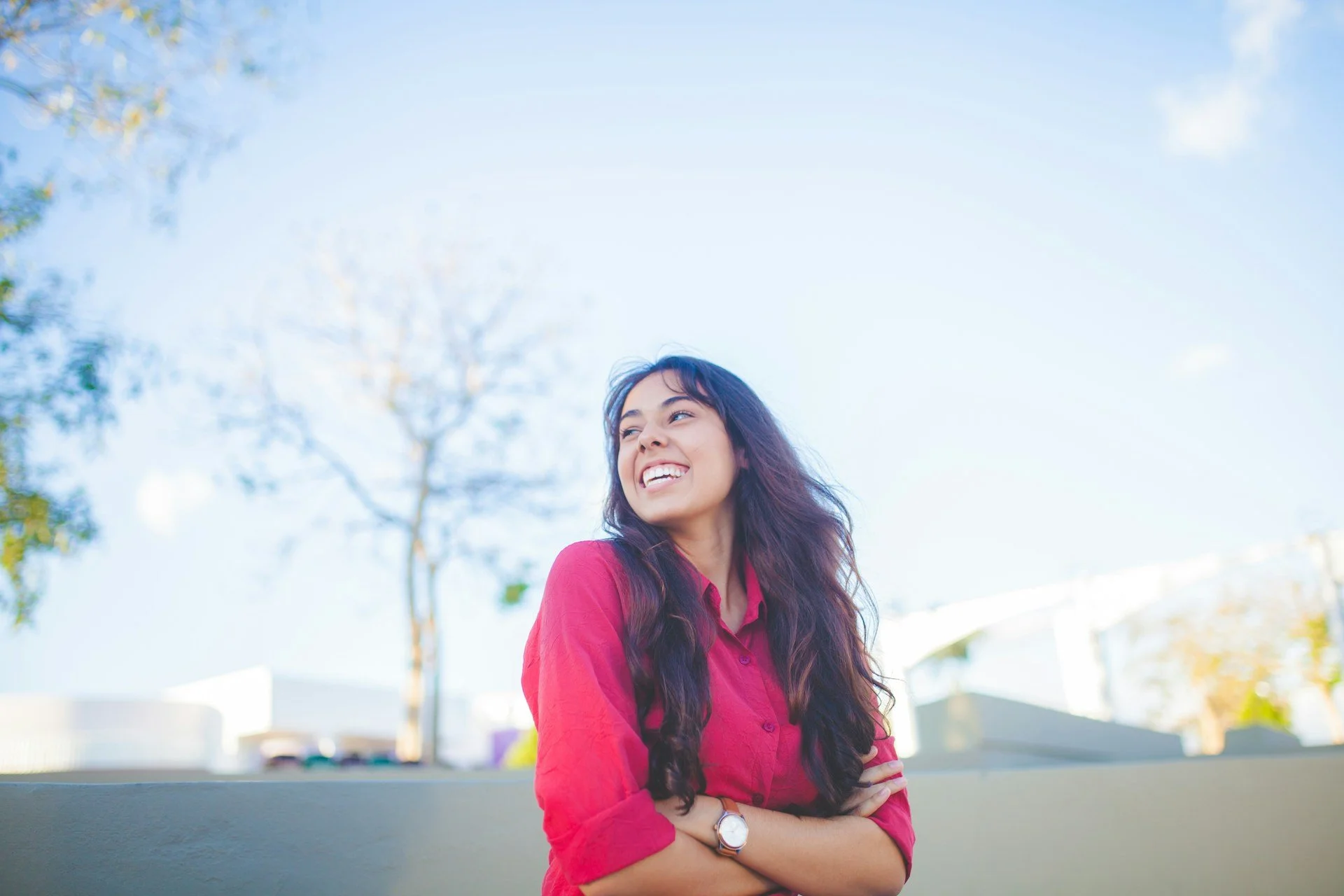 Young woman with long dark hair smiling outdoors, wearing a red shirt and a watch, standing with arms crossed in front of a blue sky and trees. A woman after trauma therapy.