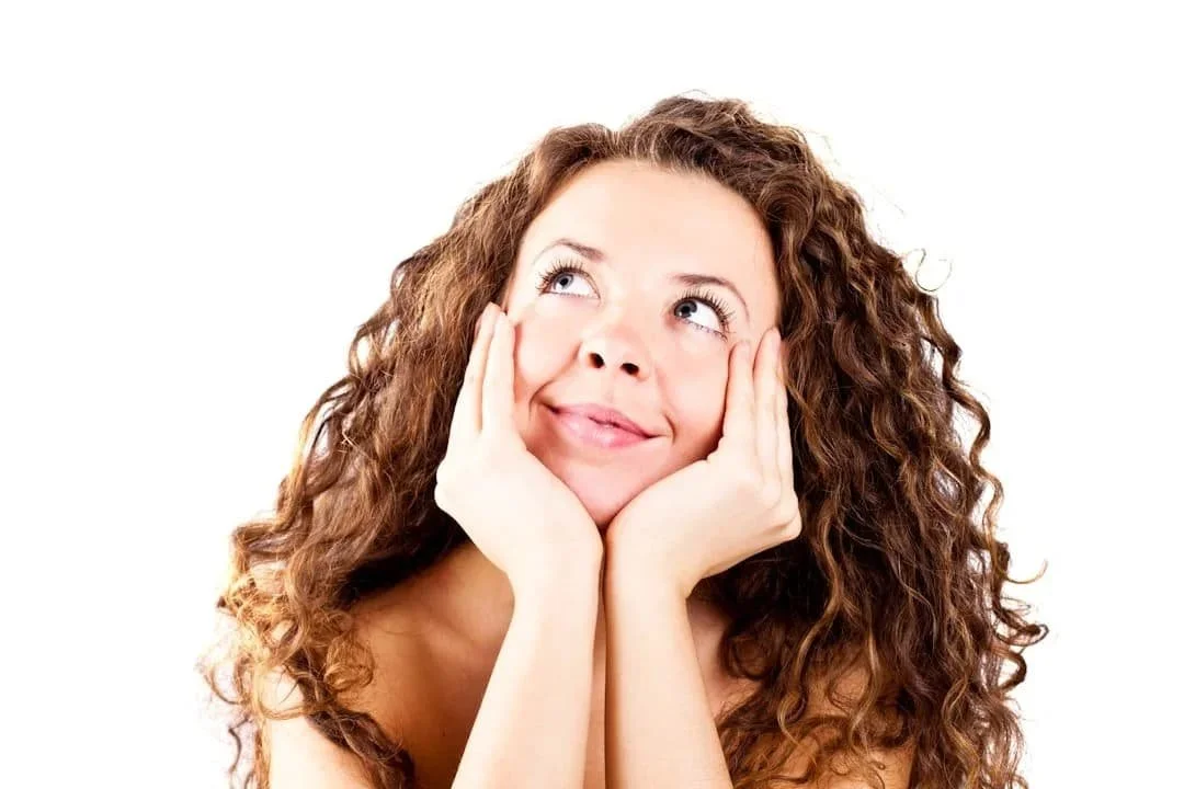Woman with curly hair resting her face in her hands and looking upward with a gentle smile, representing hope and healing through trauma therapy in Phoenix, Arizona