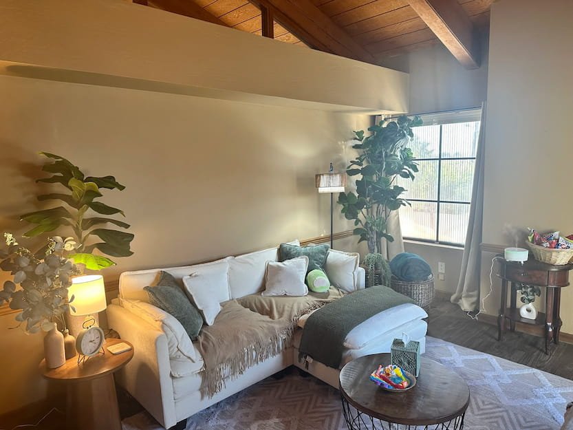 Warm therapy office with vaulted wood beam ceiling, white sectional sofa with soft pillows, large fiddle leaf fig plant, and natural light streaming through a window. Creating a grounded, safe environment for therapy sessions