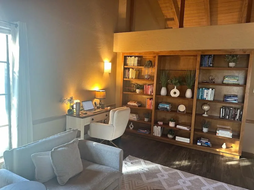 Living room with beige sofa and pillows, a white desk with a chair, bookshelf with decorative plants and books, and warm wall lighting.
