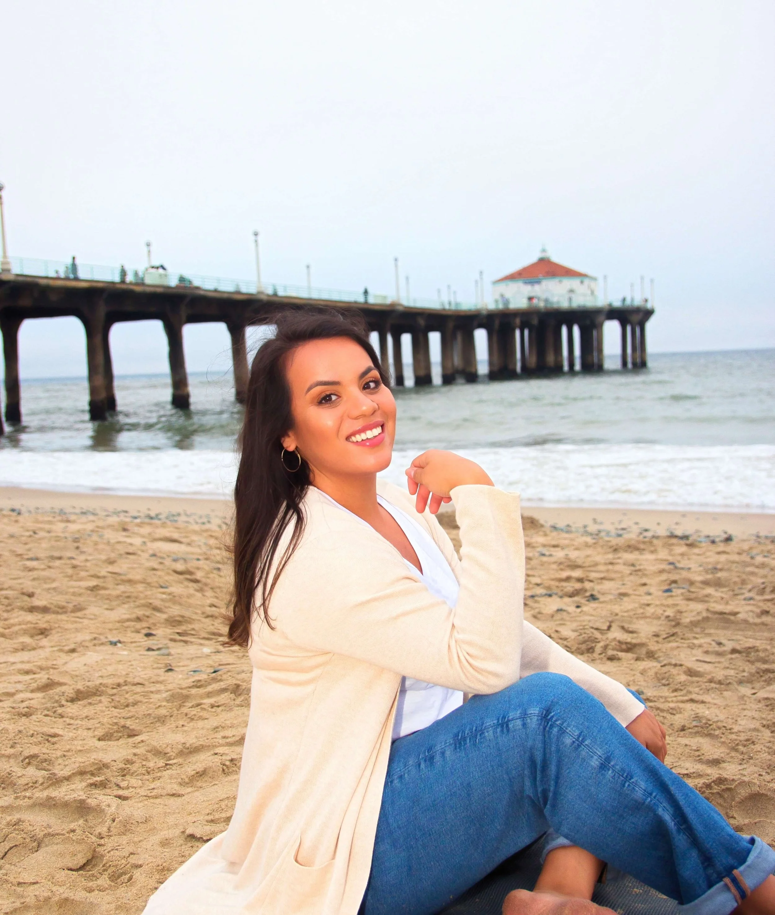 A trauma therapist sitting on the sandy beach near the ocean, with a pier and a building in the background.