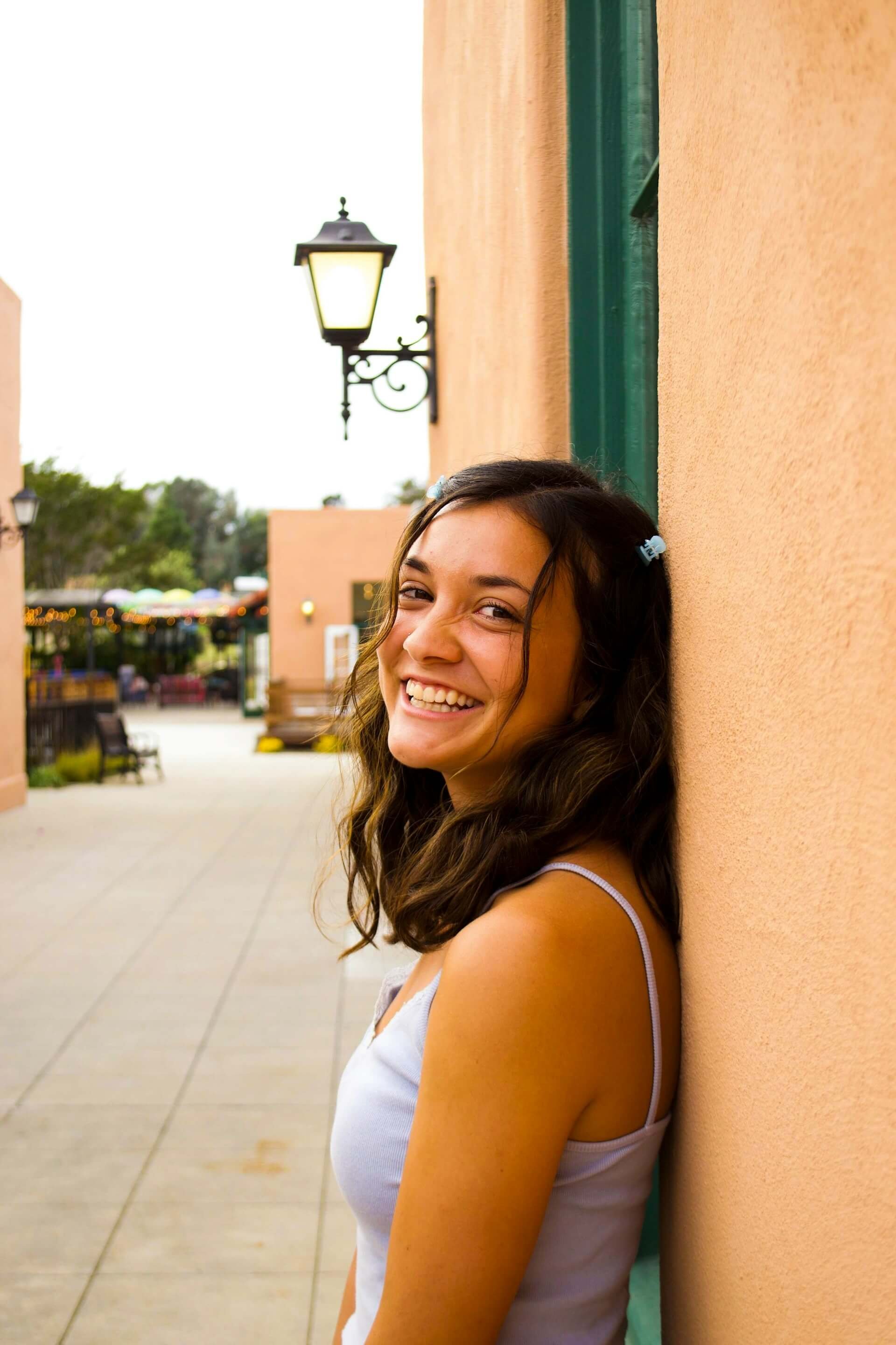 A young woman with dark brown hair smiling and leaning against an orange wall outdoors. A woman after anxiety therapy.