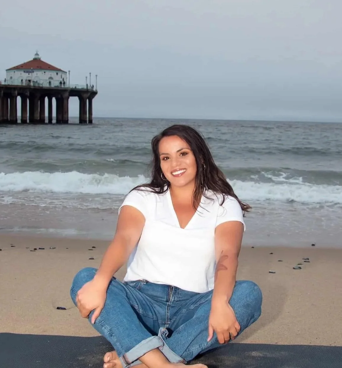 Woman sitting cross-legged on a yoga mat at the beach near a pier, smiling warmly