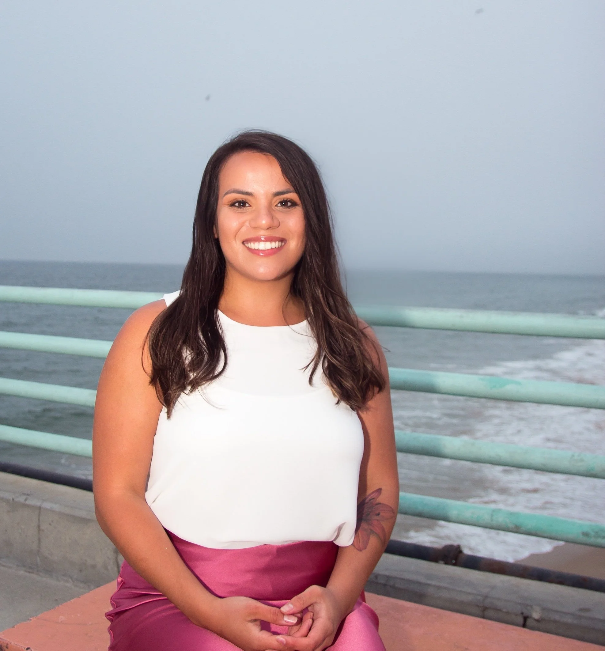 A smiling woman with long dark hair, wearing a white sleeveless top and pink pants, sitting on a wall near the ocean with a cloudy sky. A picture of trauma therapist Alexa Gonzales