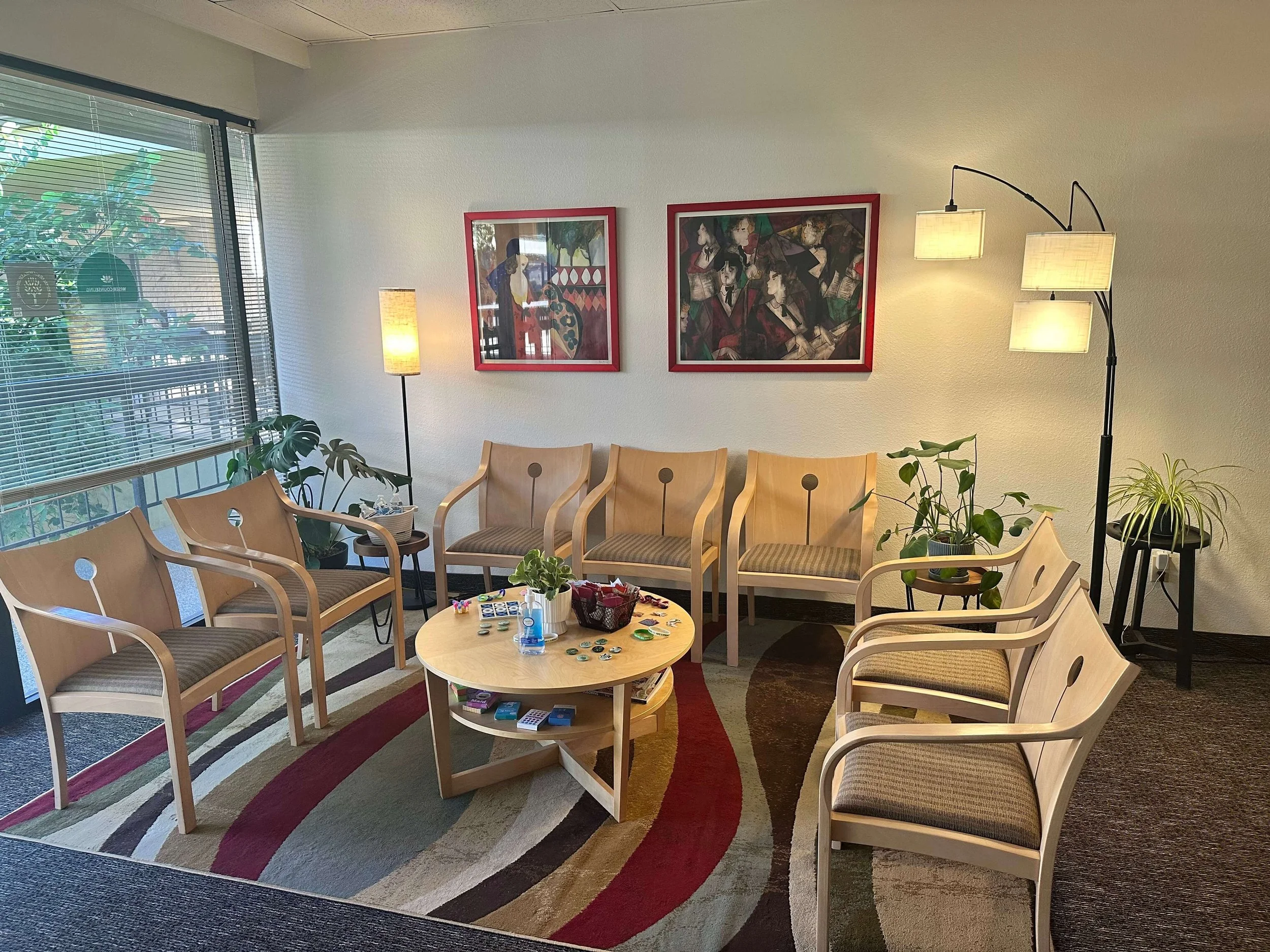 Waiting area for a Phoenix therapy office with eight wooden chairs with striped cushions arranged around a round coffee table on a colorful patterned rug. There are plants near the windows and artwork on the wall/