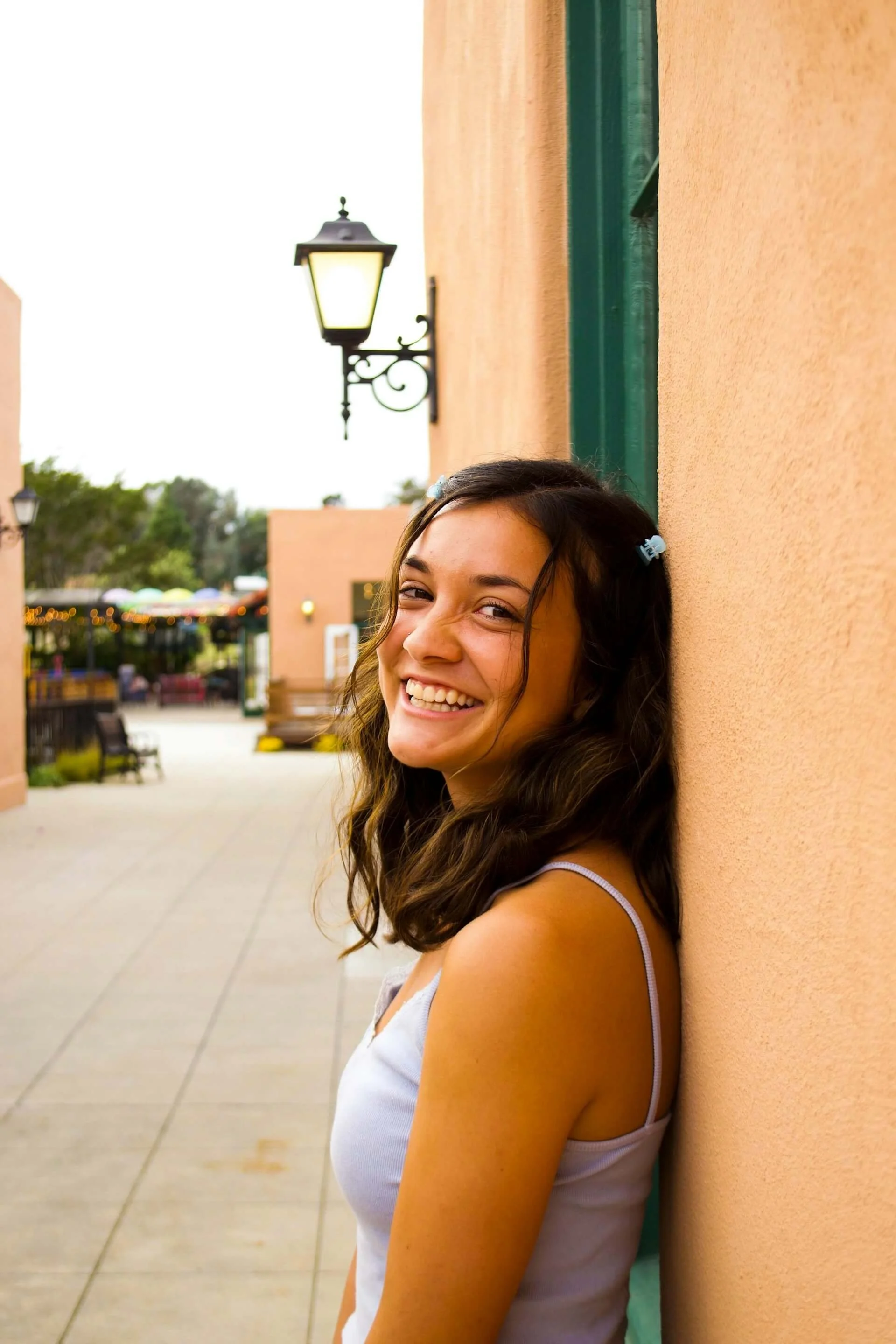 A young woman with wavy brown hair smiling after anxiety therapy and leaning against a peach-colored wall outdoors at sunset.