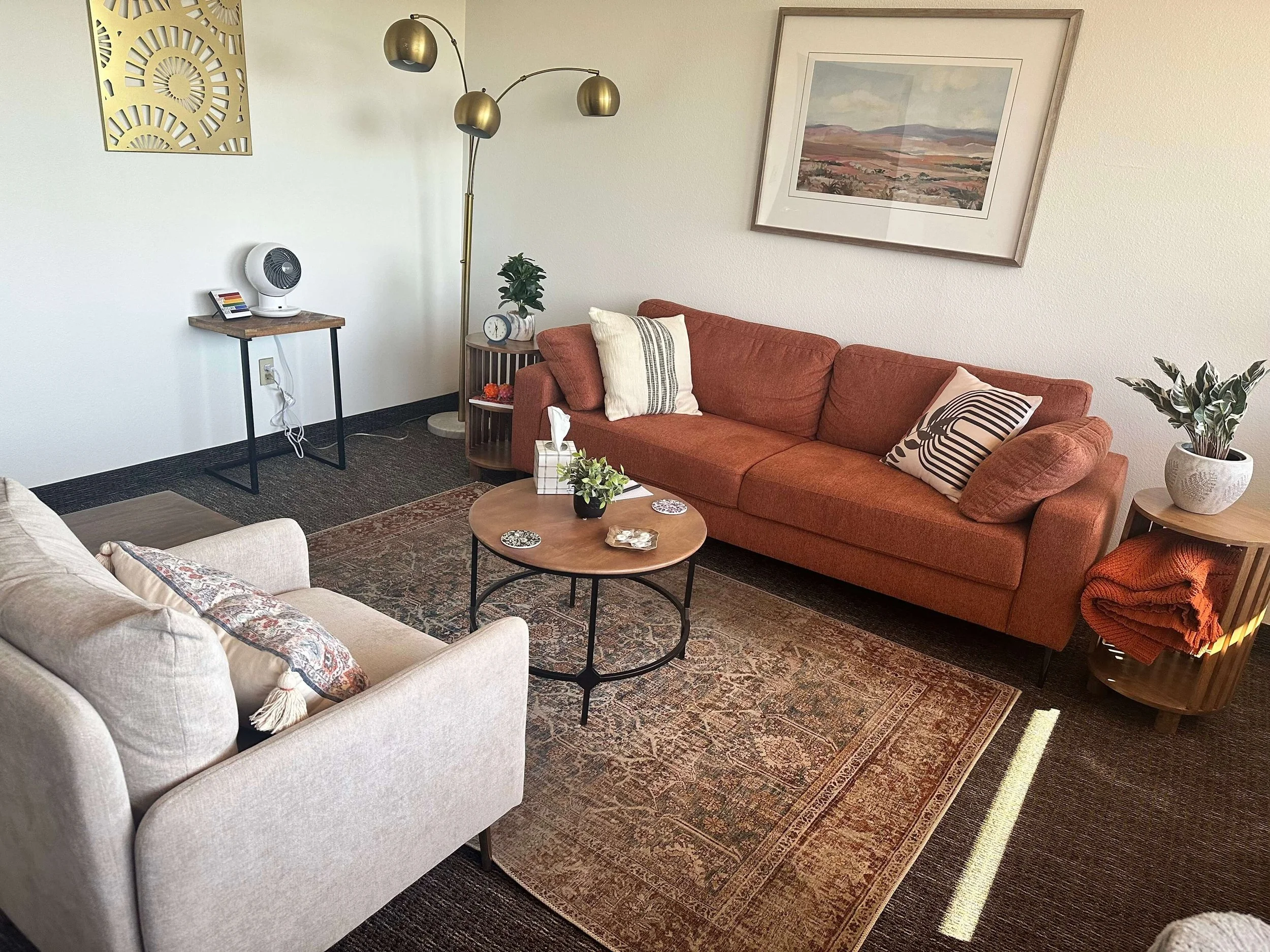 Phoenix Therapist office with beige and rust-colored sofas, a round wooden coffee table, potted plants, a framed landscape painting, a standing lamp with multiple gold-colored shades, a side table with a clock.