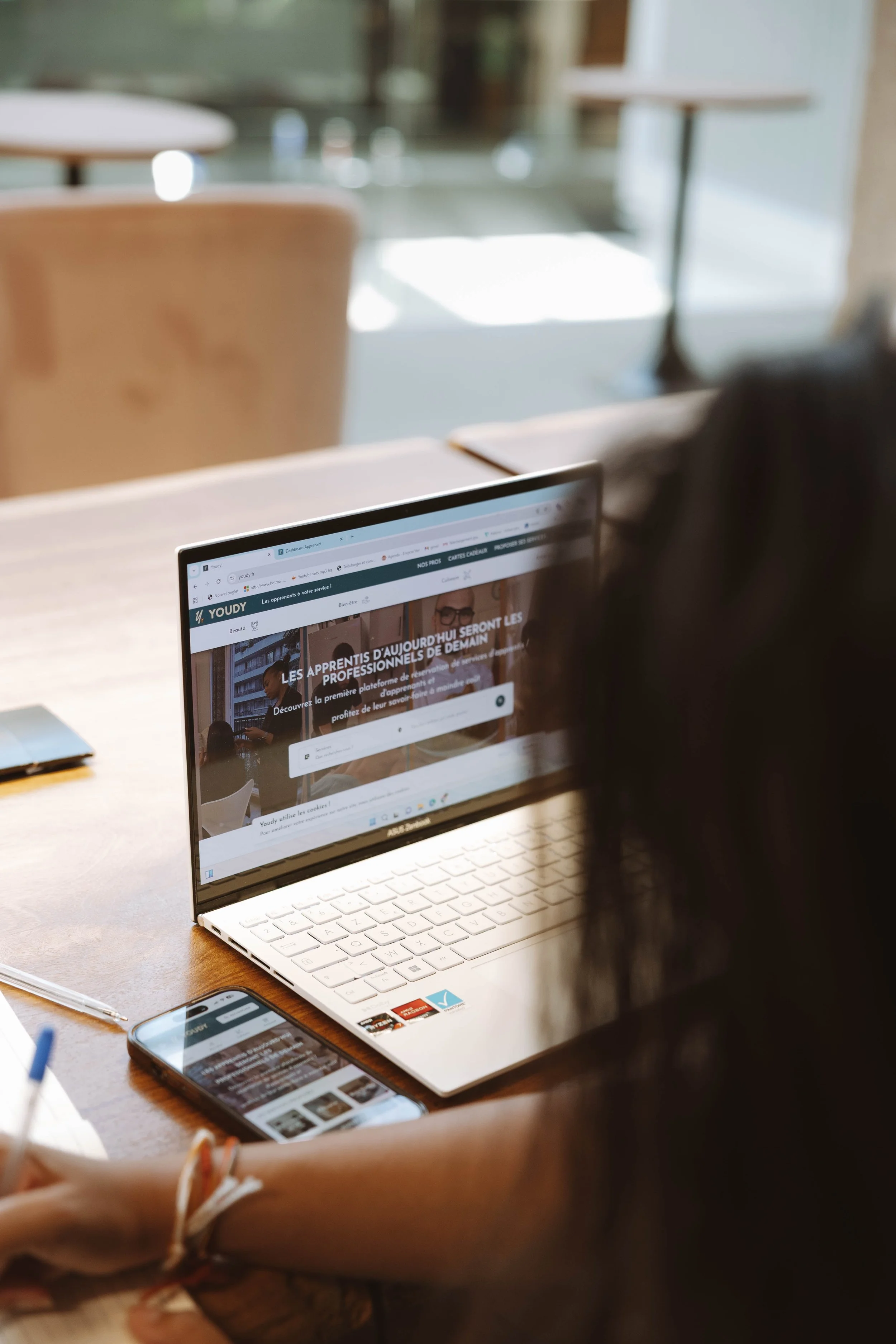 Une personne regarde un ordinateur portable sur une table en bois, affichant une page web en français. Un téléphone est aussi posé sur la table à côté.