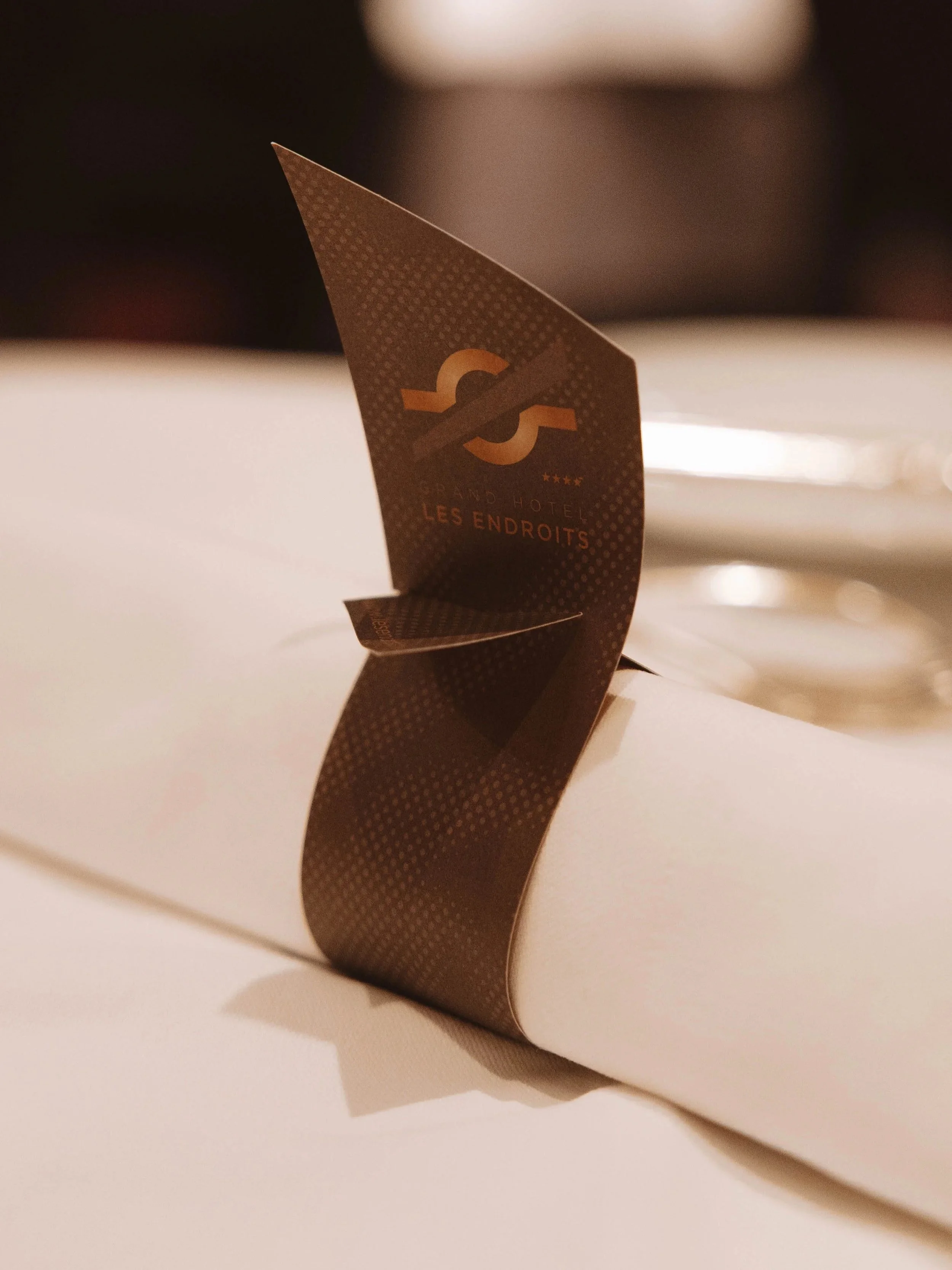Marque-page ou marqueur de table pour le Grand Hôtel Les Endroits, inséré dans une nappe blanche sur une table de restaurant.