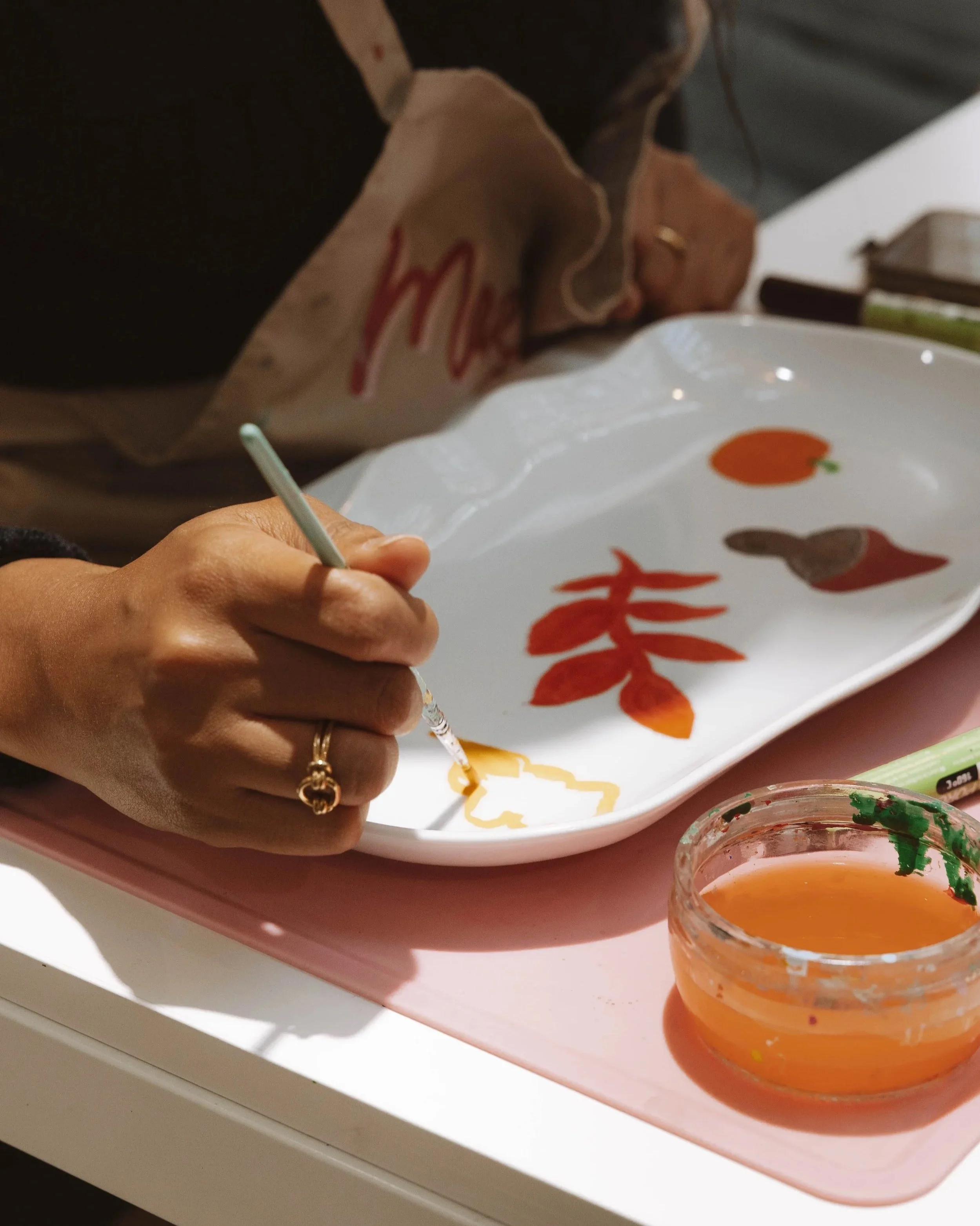 Une personne peint des motifs de couleur jaune sur une assiette blanche en céramique, avec un petit pot de peinture à côté et d'autres dessins en rouge et marron sur la vaisselle.