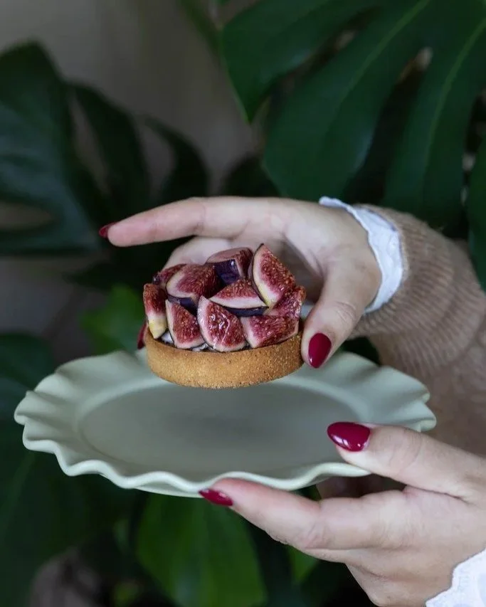 Une personne tient une tartelette aux figues fraîchement préparée, avec une crème ou un garniture en-dessous, sur une assiette blanche. La personne a les ongles peints en rouge et porte un pull beige et une chemise blanche.