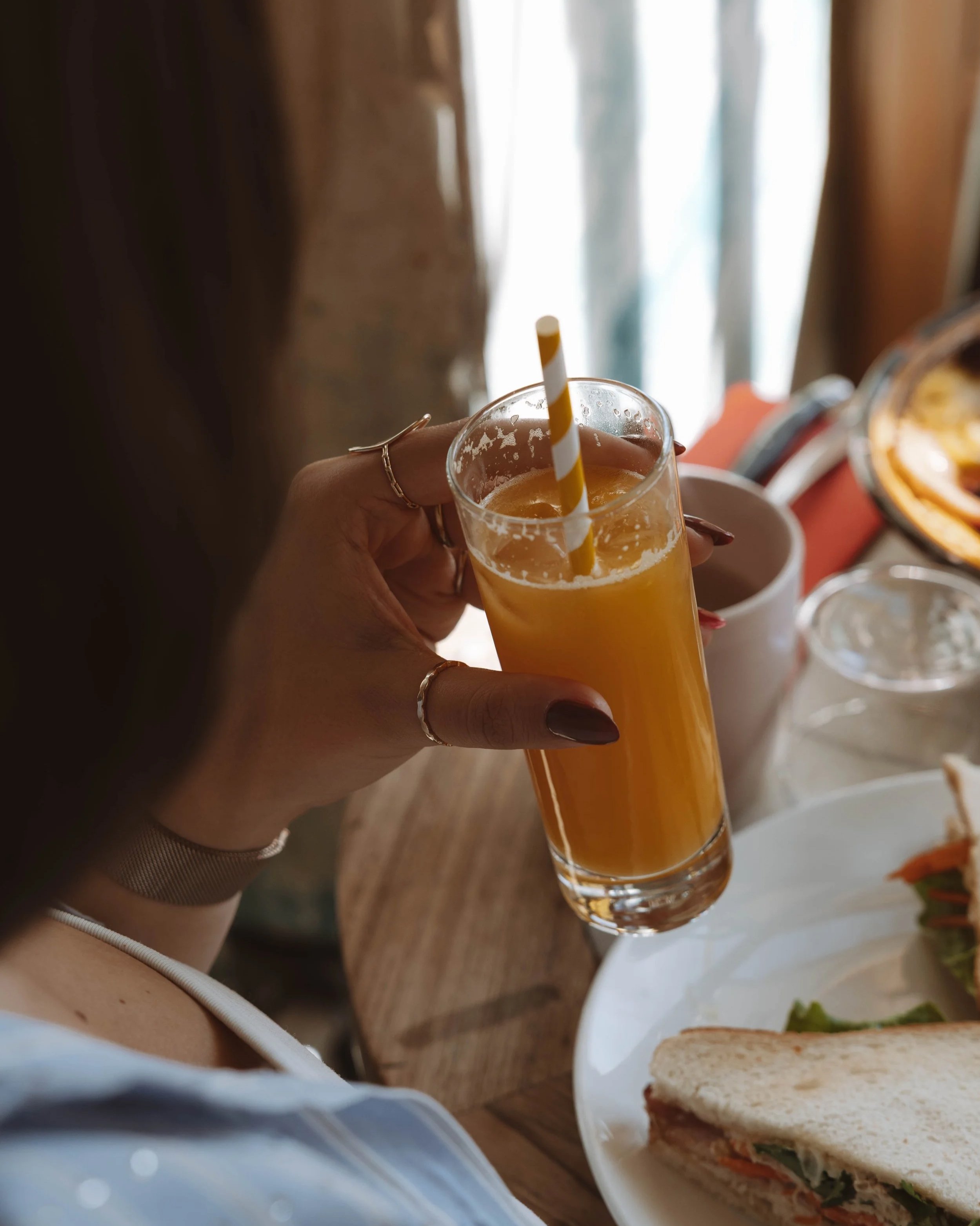 Une personne tient un verre de jus d'orange avec une paille jaune et blanche, avec un plateau de nourriture à côté, y compris un sandwich au pain blanc et une tasse. Il y a de la lumière naturelle en arrière-plan.