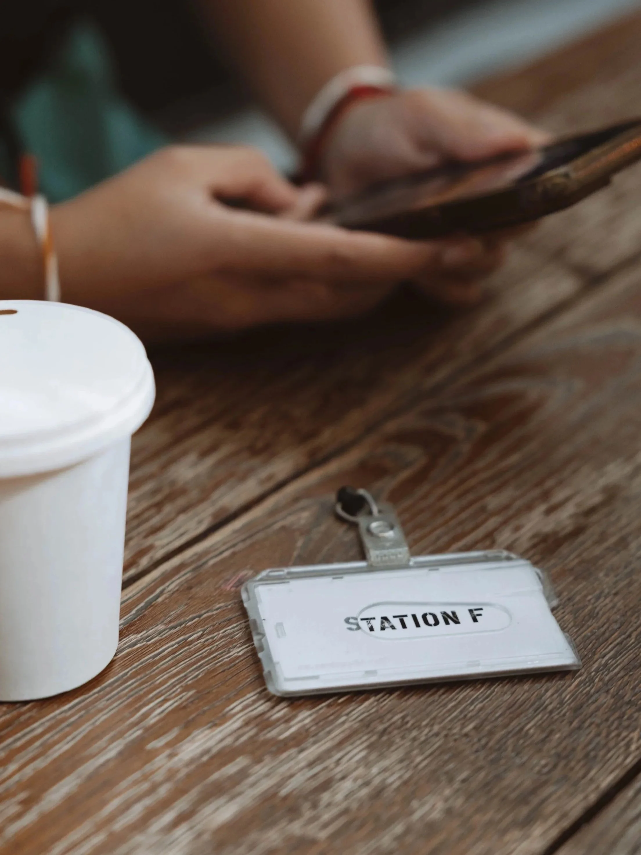 Une personne tenant un téléphone courtisée, une tasse blanche sur une table en bois, et un badge avec l'inscription "STATION F" posé sur la table.