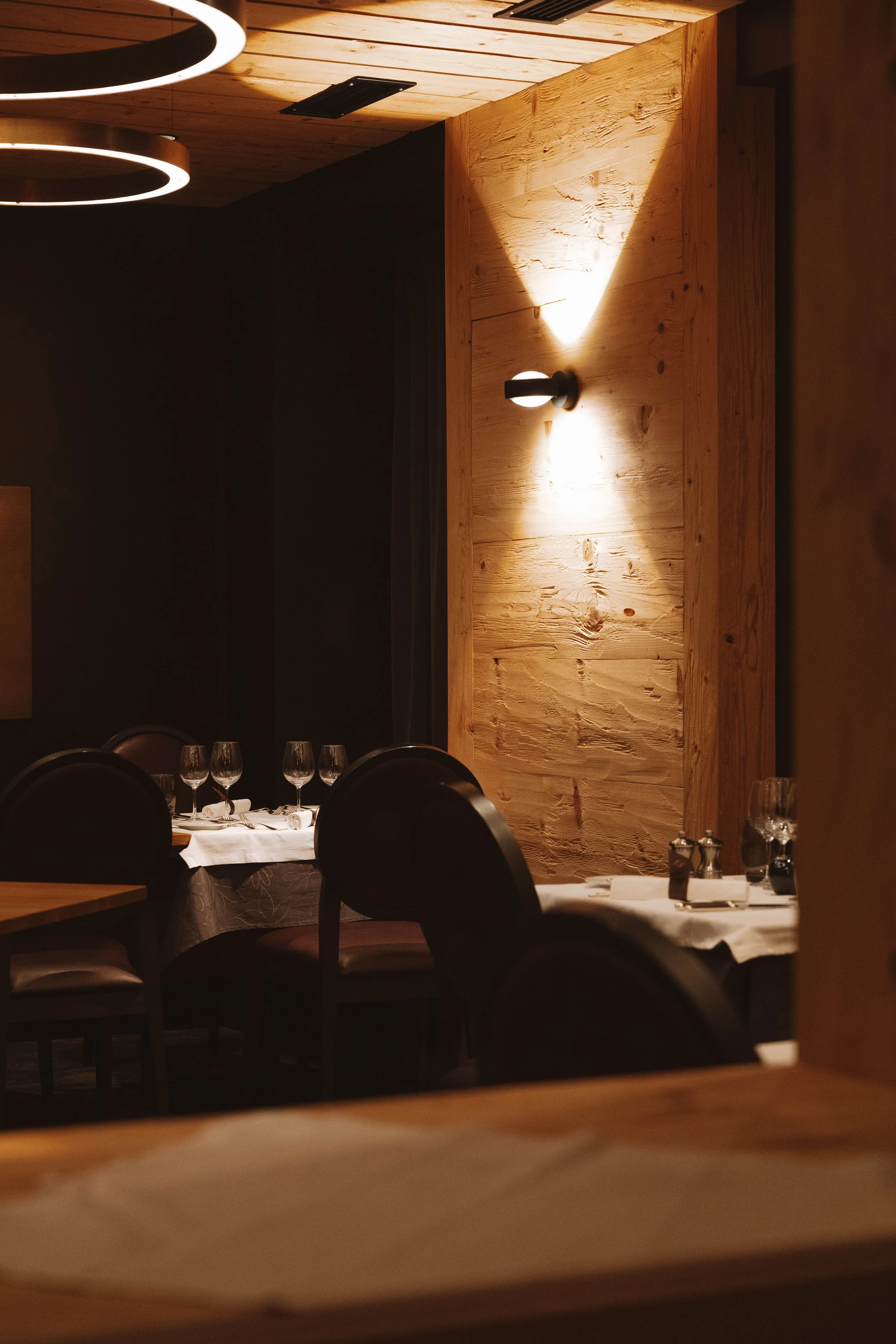 Salle à manger élégante avec tables dressées, verres à vin et assiettes, éclairée par un éclairage tamisé, avec un mur en bois et un éclairage mural en fond.