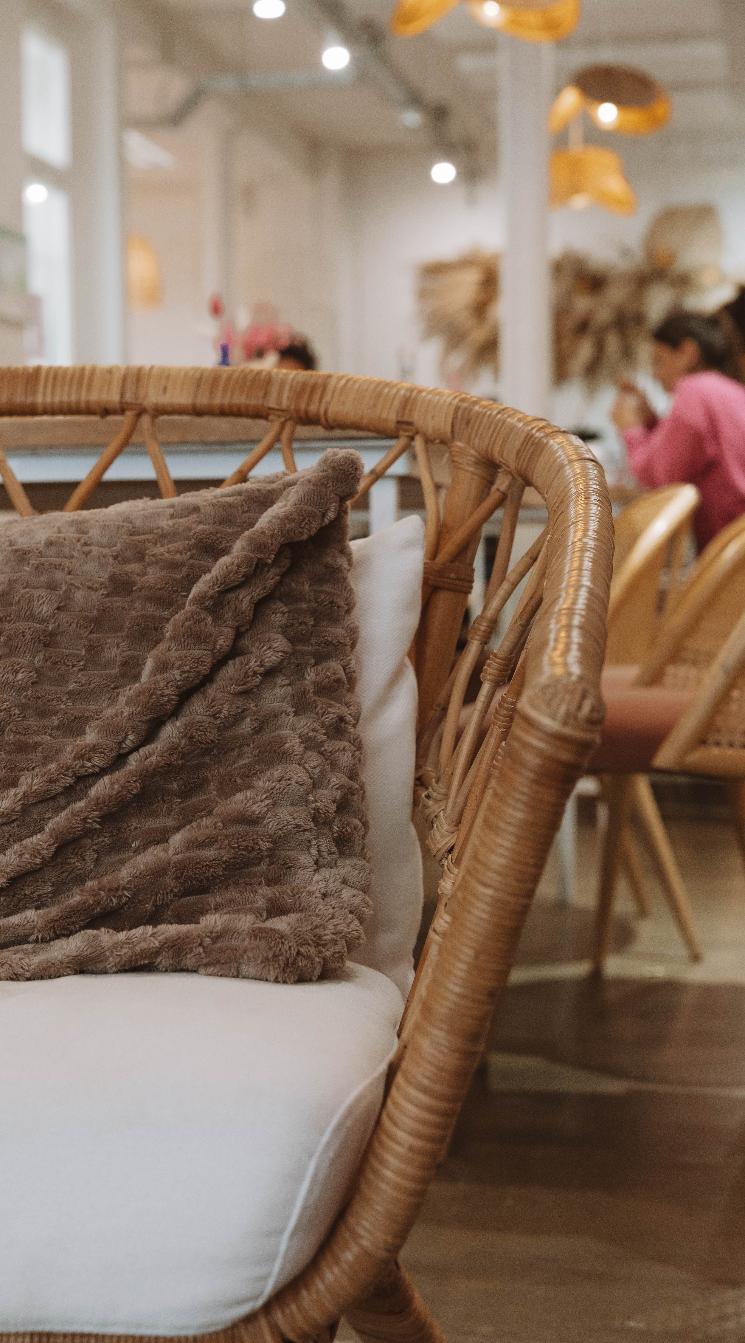 Intérieur d’un café ou salon de thé, avec un fauteuil en rotin, coussin en molleton marron et blanc, et des personnes assises en arrière-plan.