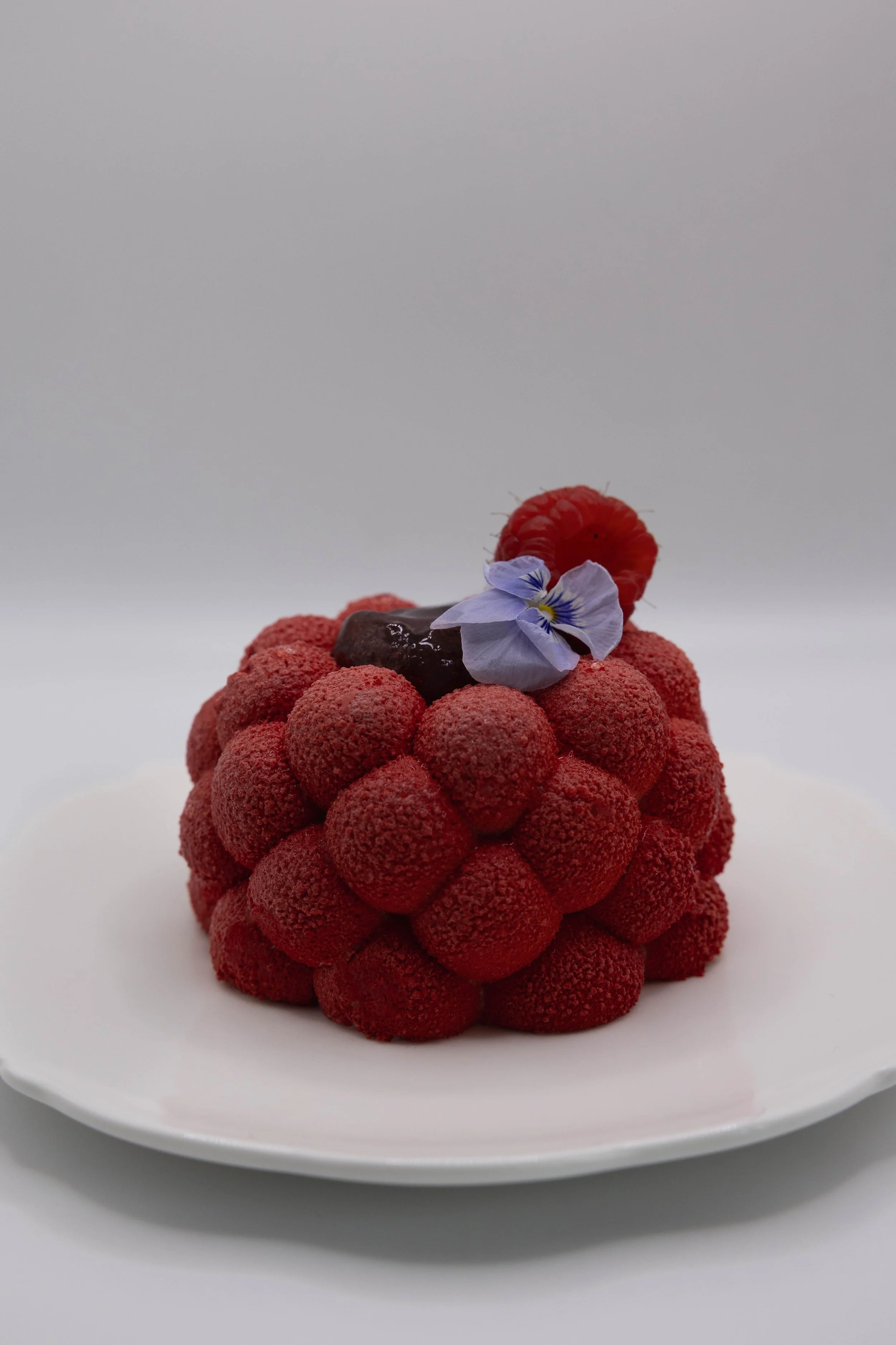 Gâteau décoré avec des framboises, une fleur violette, et une petite mûre, présenté sur une assiette blanche.