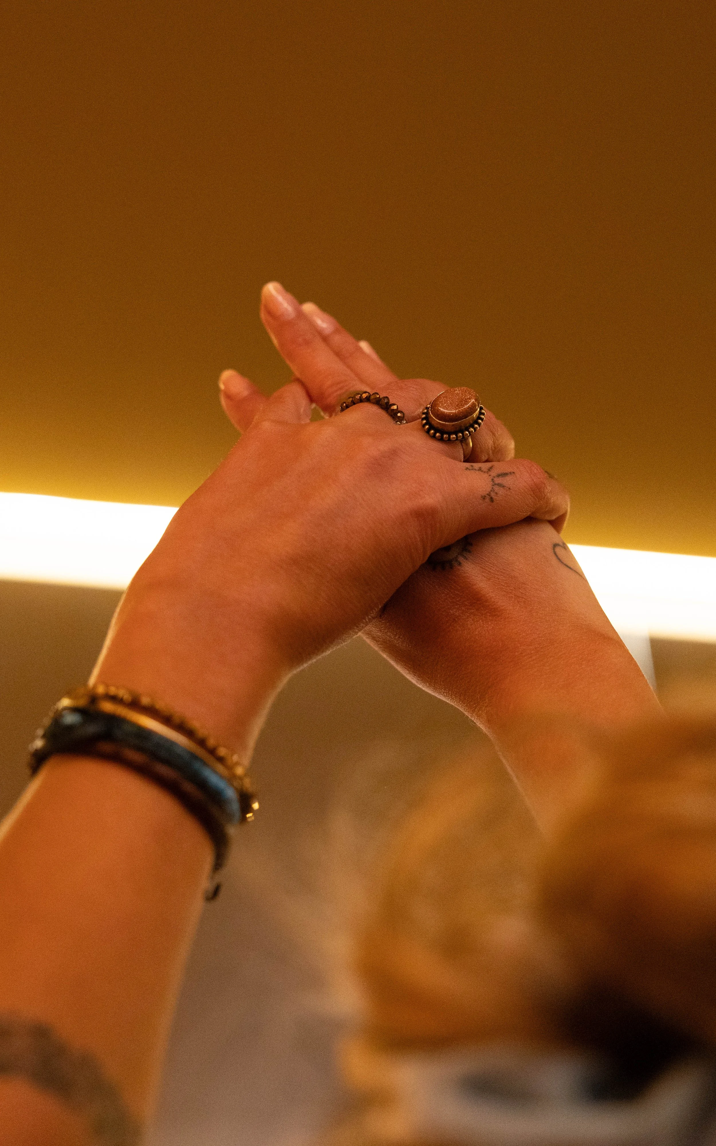 Deux mains entrelacées avec des bijoux, tenant légèrement une autre main, contre un fond lumineux et une surface sombre.