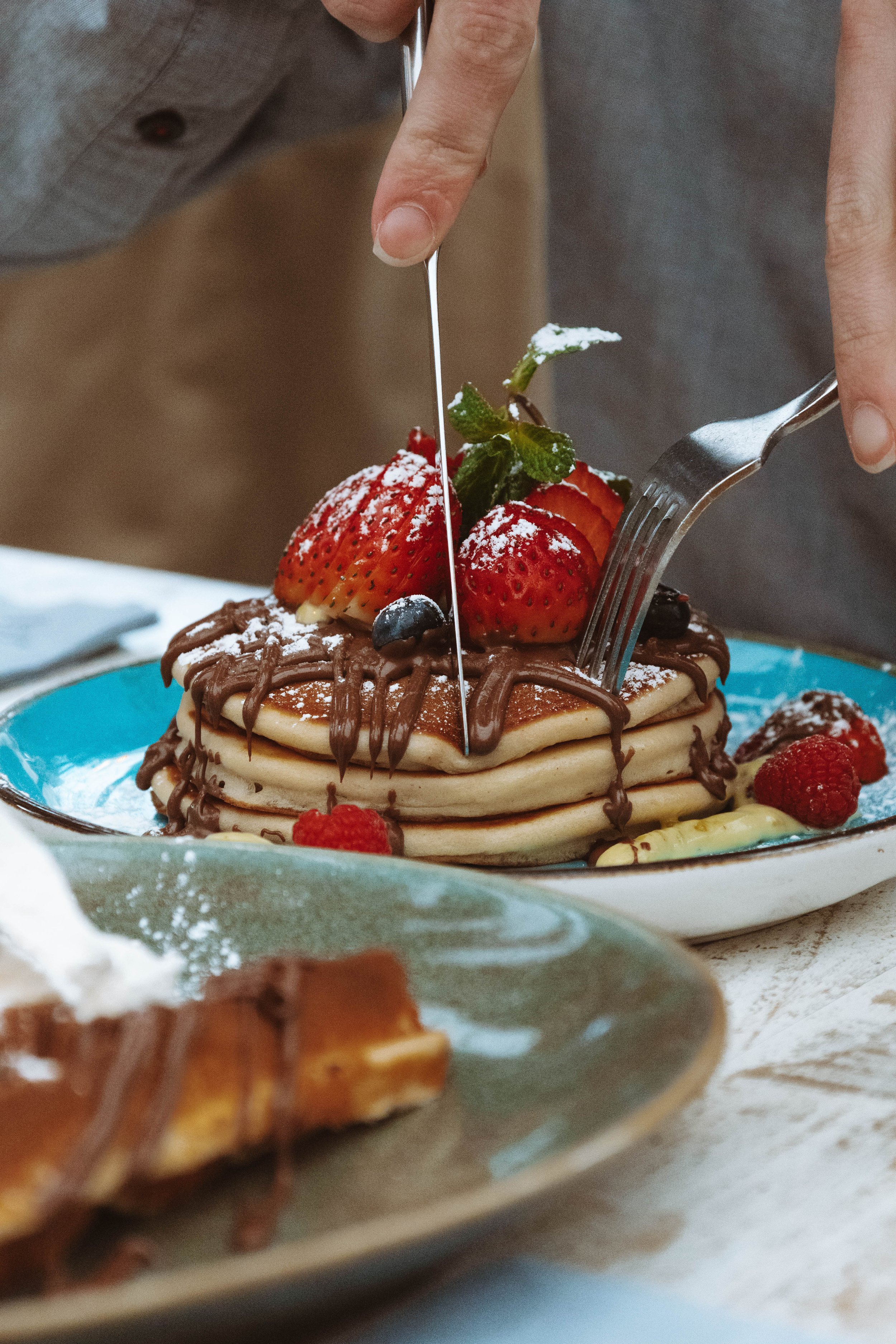 Des pancakes garnis de fruits rouges, de chocolat fondu, et de feuilles de menthe, avec une personne en train de les couper avec une fourchette et un couteau.