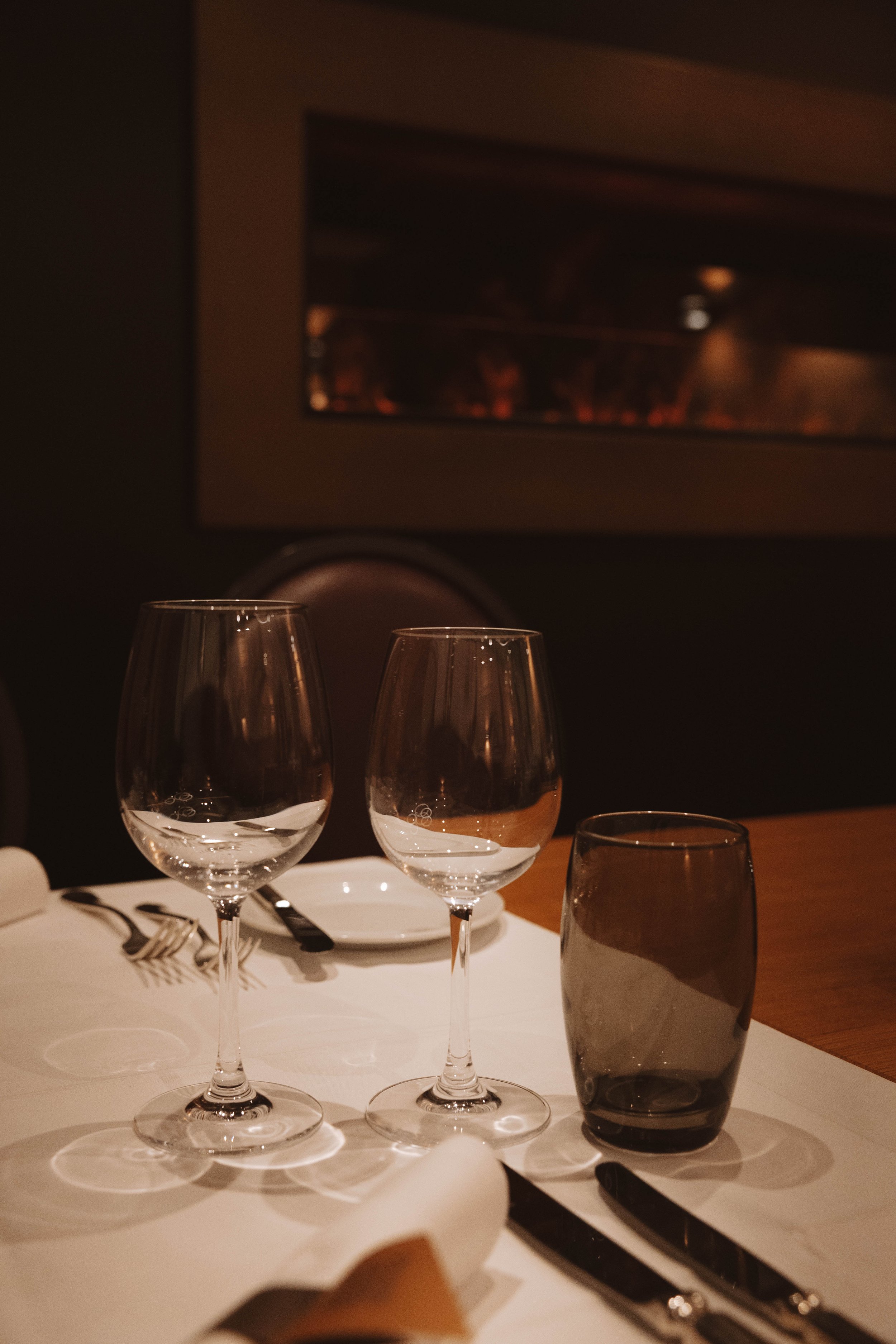 Deux verres à vin vides, un verre teinté foncé, une assiette blanche, des fourchettes et couteaux en métal sur une nappe blanche, dans un restaurant avec ambiance chaleureuse.