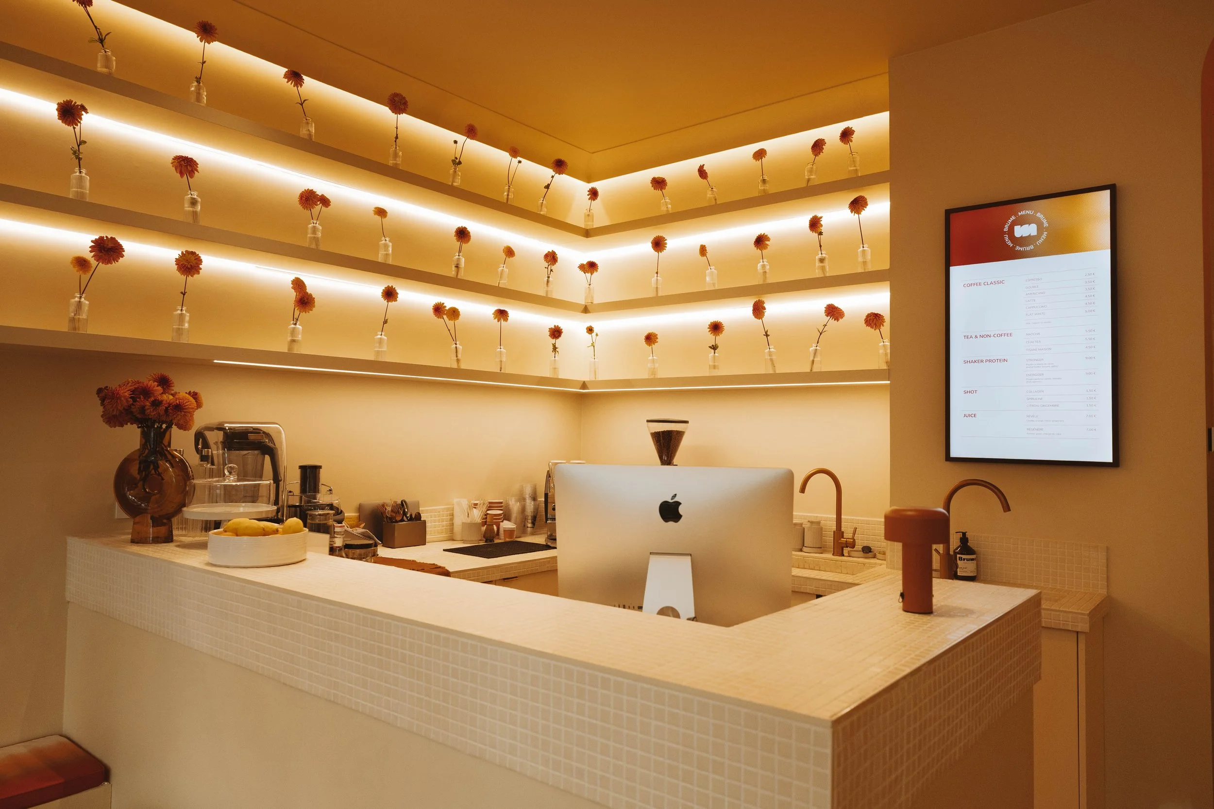 Un comptoir de café élégamment décoré avec des fleurs orange dans des vases, une affiche numérique affichant le menu, et un ordinateur Apple sur le comptoir.