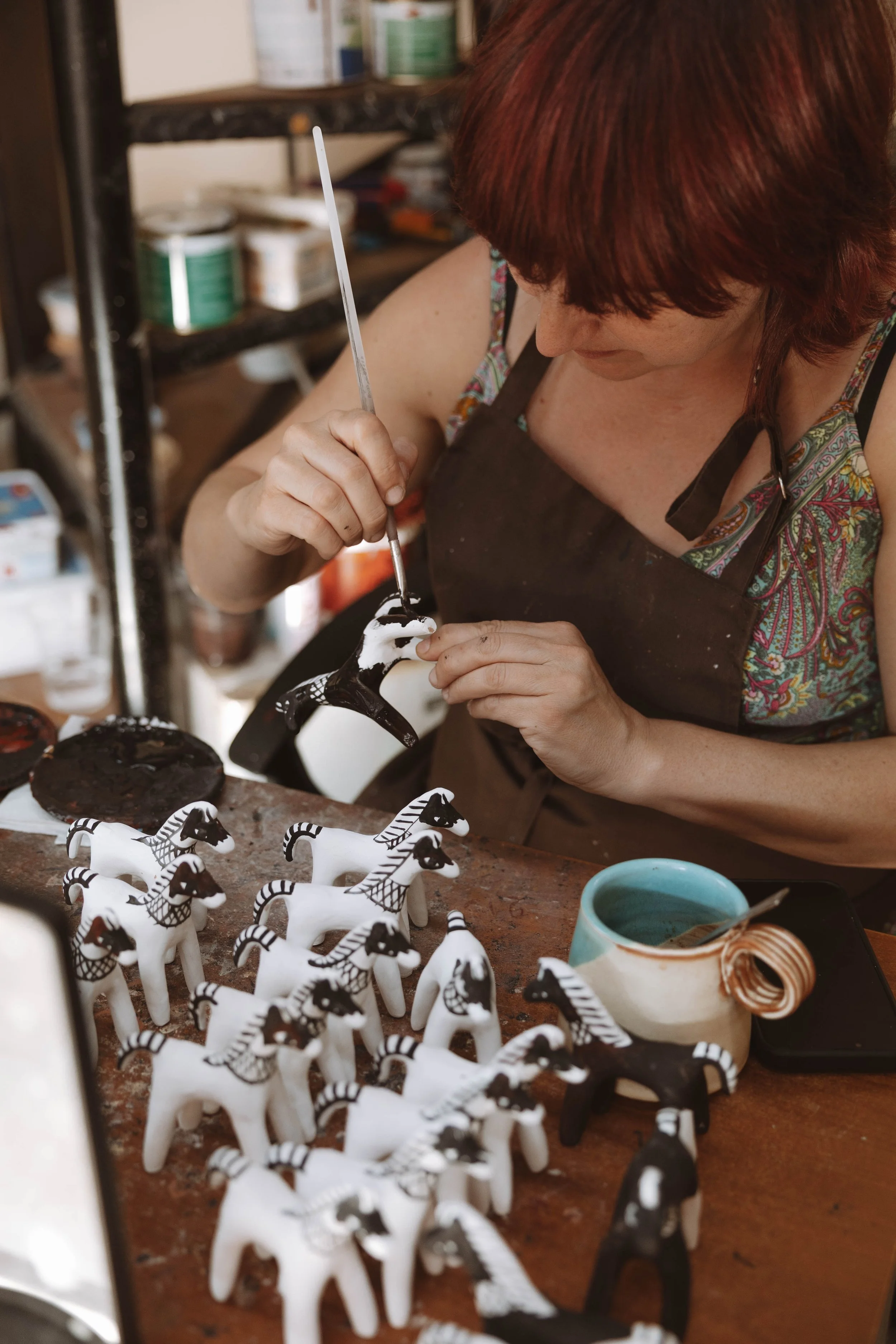 Une femme peignant une figurine en forme de cheval, entourée de plusieurs autres figurines de chevaux blancs et noirs sur une table, avec des pinceaux et des outils de peinture.