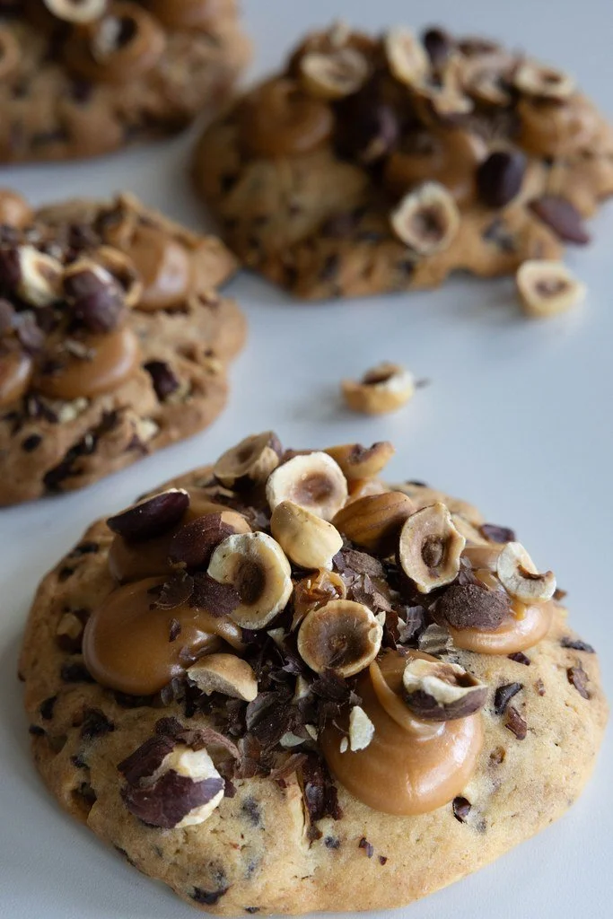 Cookies avec des noix de macadamia, des morceaux de chocolat et du caramel sur une surface blanche.