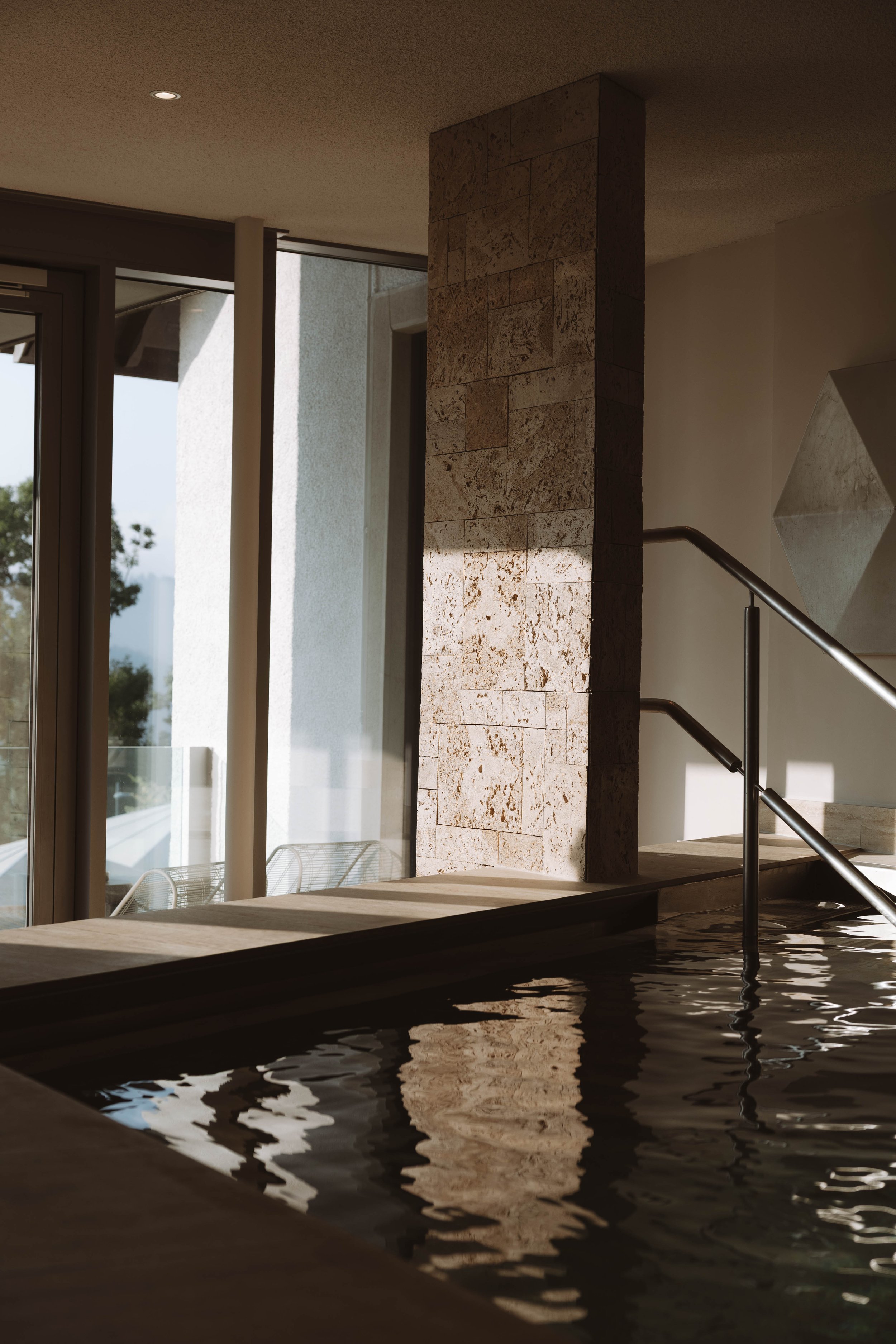 Intérieur d'une piscine intérieure avec une paroi en pierre, une rampe en métal et de grandes fenêtres laissant entrer la lumière naturelle.