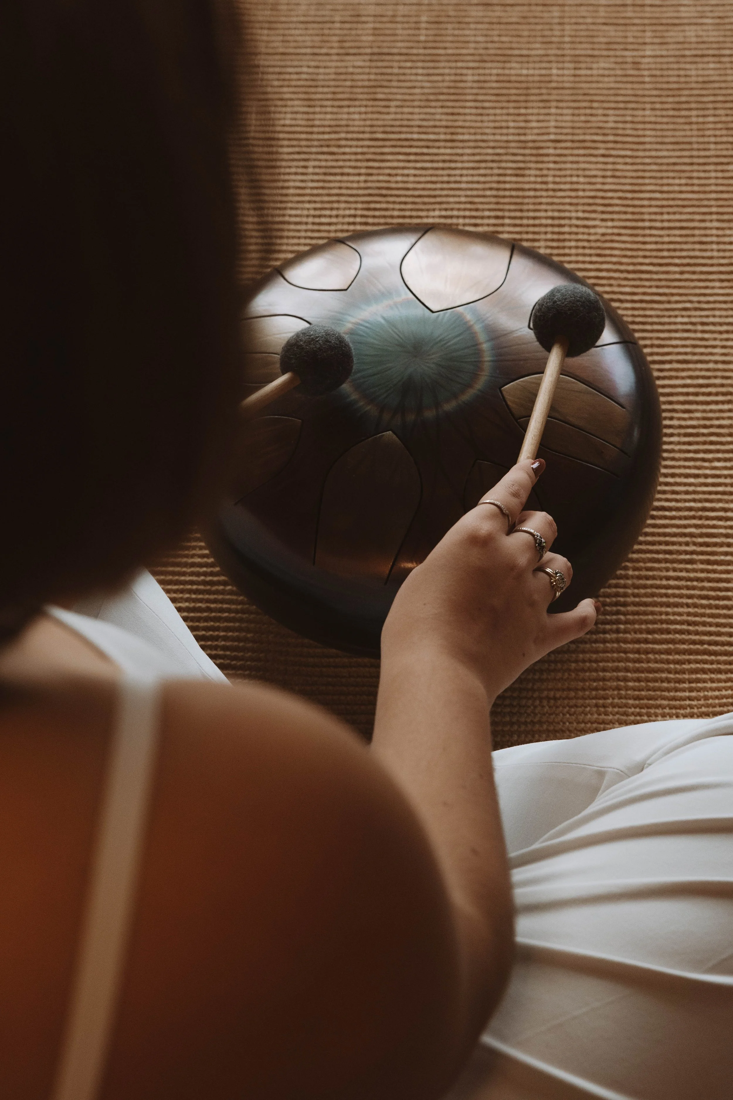 Personne jouant du tambour de vapeur de style tibétain, vue de dessus, assise sur un tapis beige, portant des vêtements blancs et des bijoux.