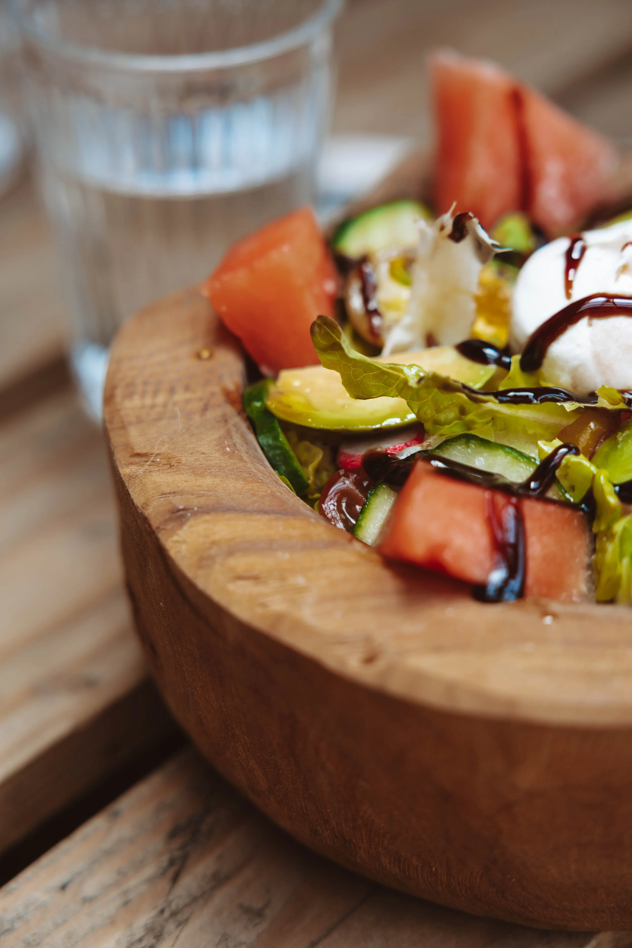 Salade de légumes frais avec morceaux de melon, avocat, concombre, radis, laitue, garnie de sauce balsamique, servie dans un bol en bois, avec un verre d'eau en arrière-plan.