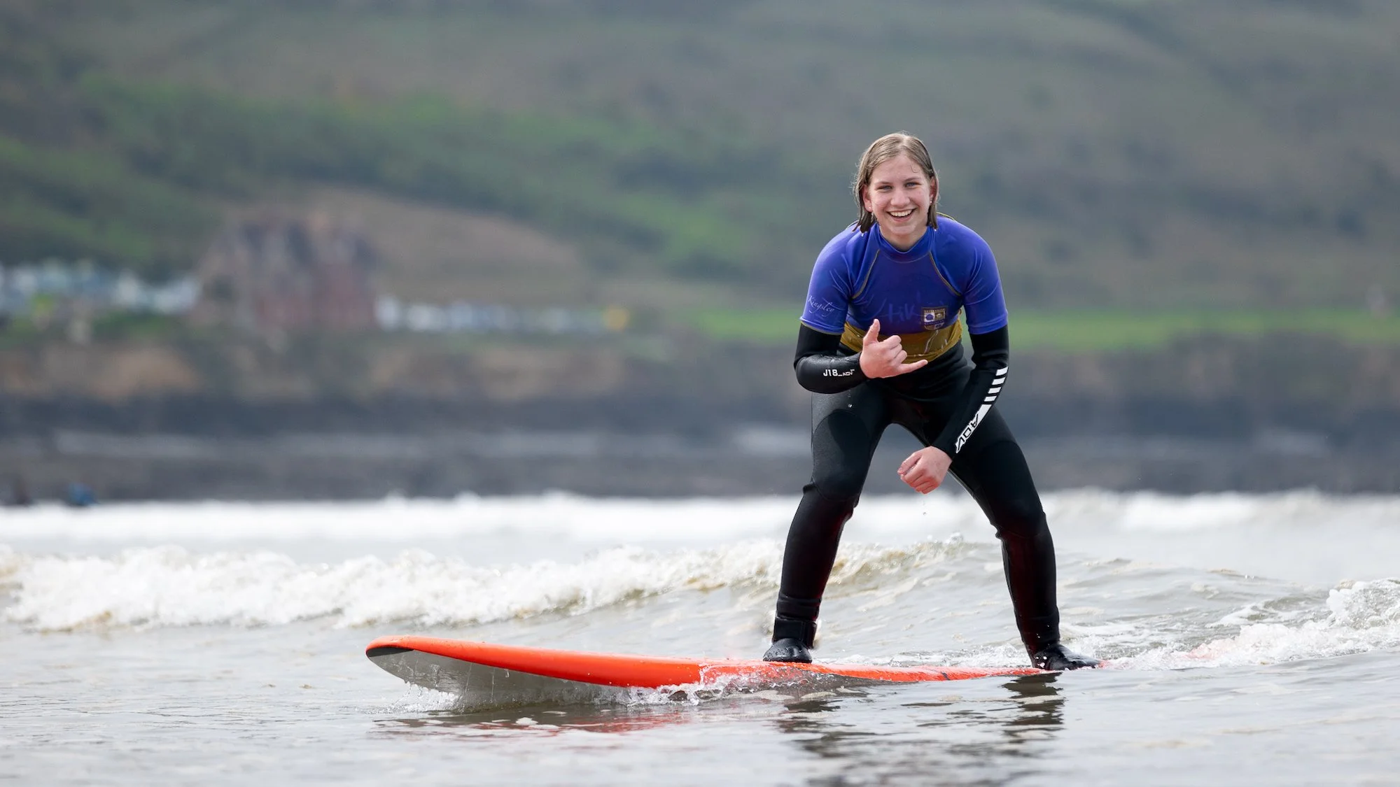 Joven sonriendo mientras practica surf en la playa con un fondo de colinas verdes y edificios