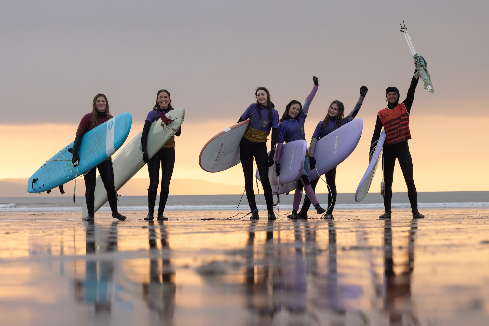 Grupo de seis personas en la playa con tablas de surf durante el atardecer, sonriendo y celebrando.