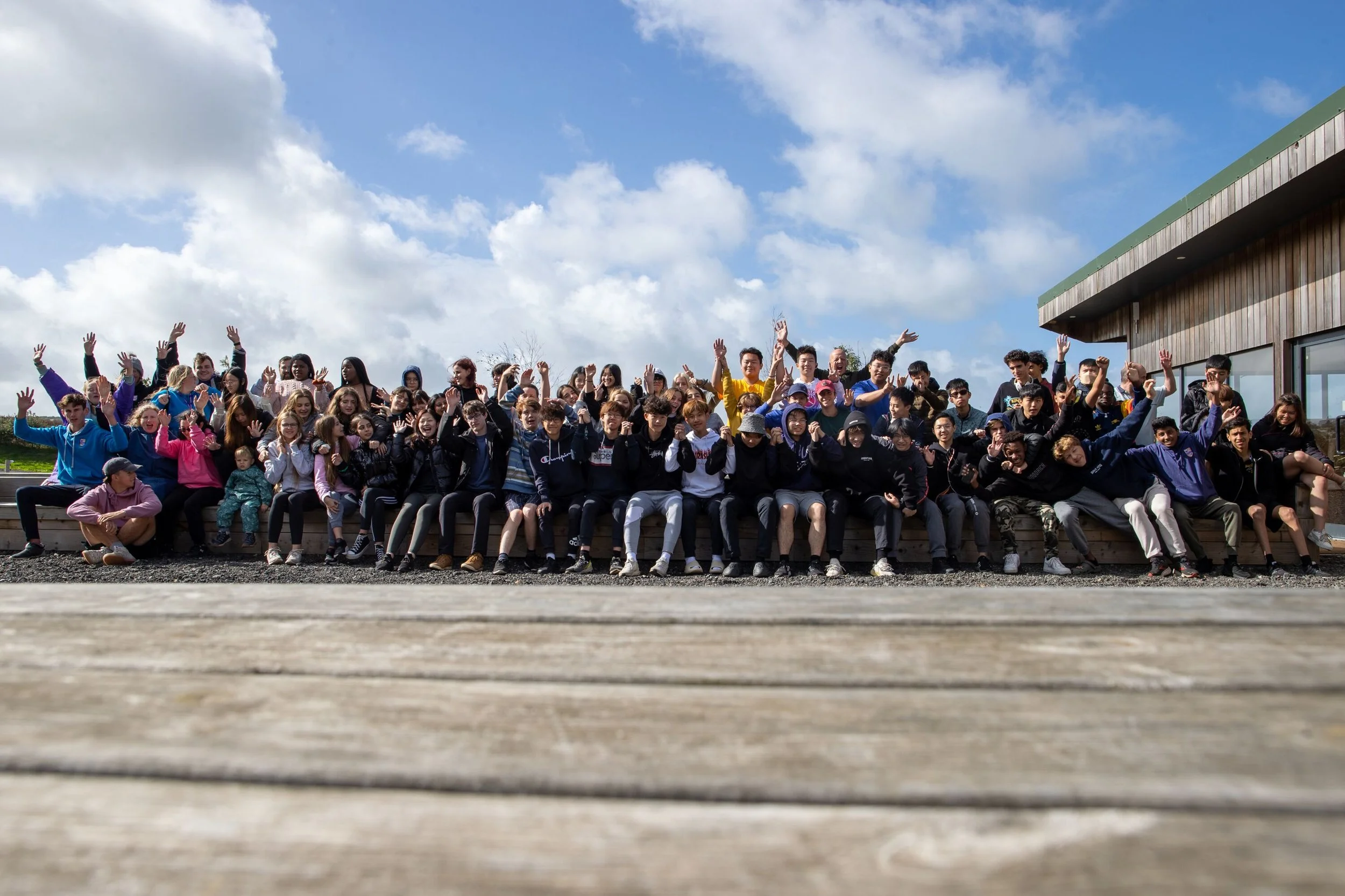 Grupo grande de jóvenes y niños en una fila, algunos sentados y otros de pie, en un espacio exterior con cielo nublado, todos saludando y sonriendo, con un edificio de madera y ventanas a la derecha.