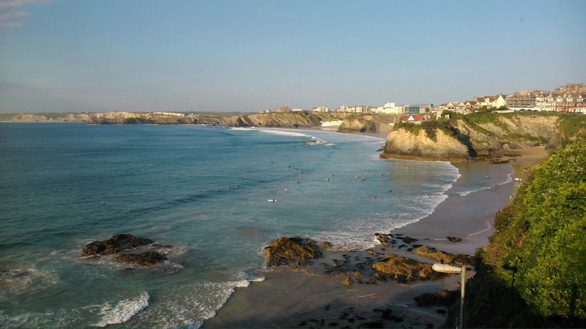 Vista panorámica de una playa con rocas, olas y varias personas practicando surf, con casas y edificios en los acantilados al fondo y un cielo despejado.