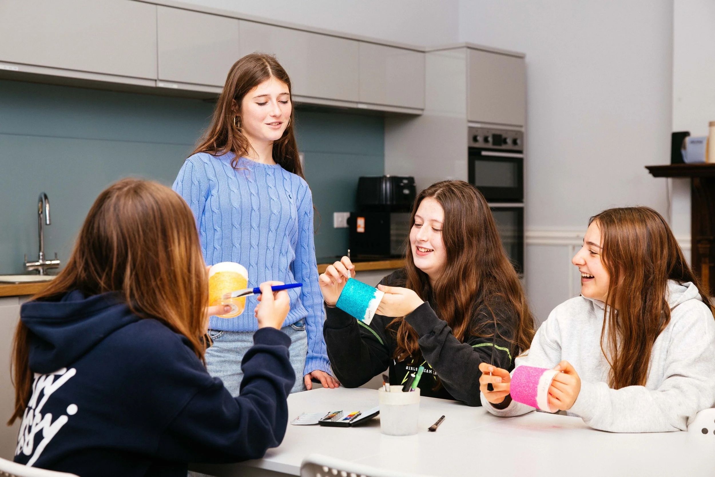Cuatro chicas en una mesa de cocina haciendo manualidades con pintura y cinta adhesiva colorida, mientras una otra les muestra algo y están sonriendo.