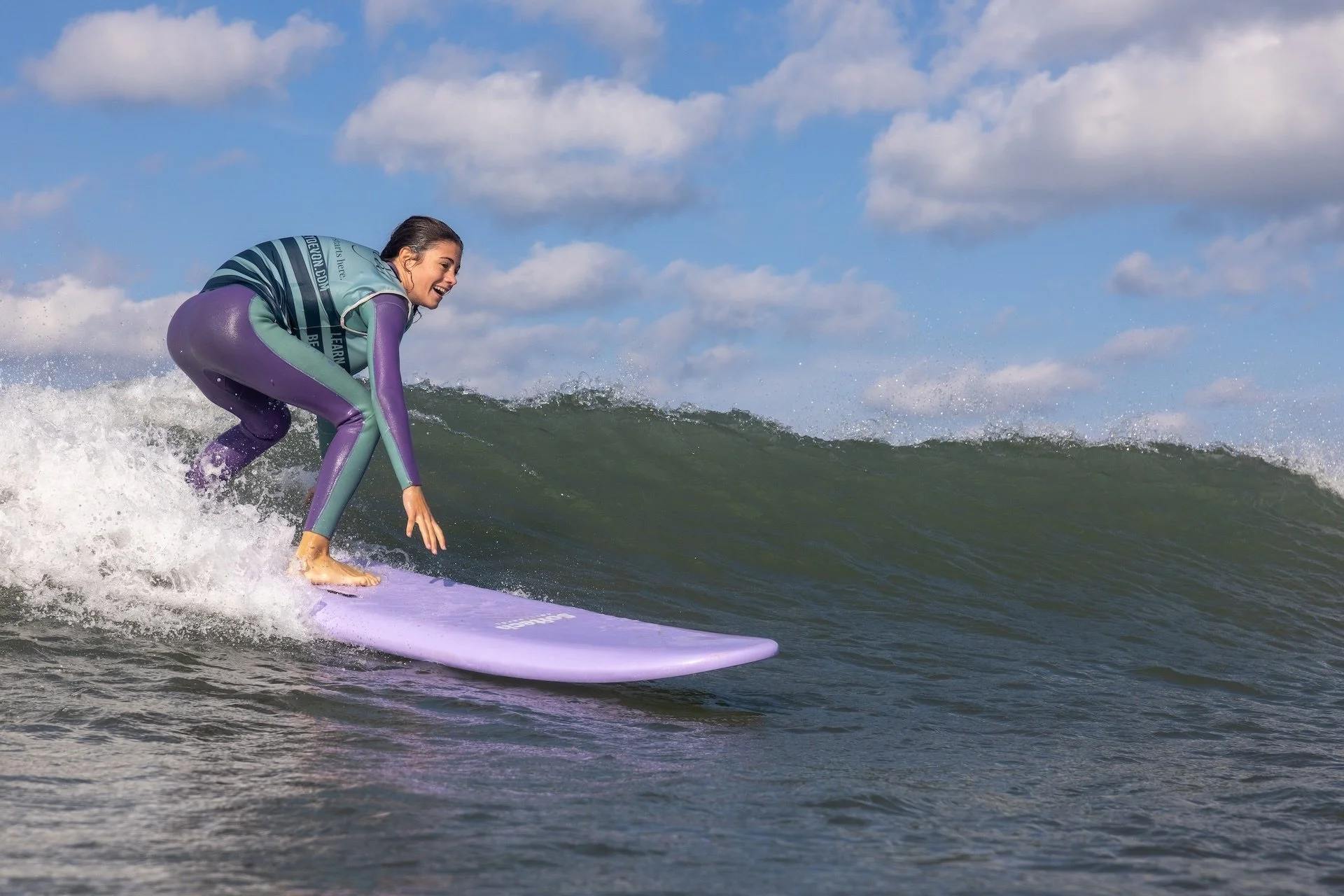 Mujer surfeando en el océano sobre una tabla morada, con un cielo despejado y algunas nubes.
