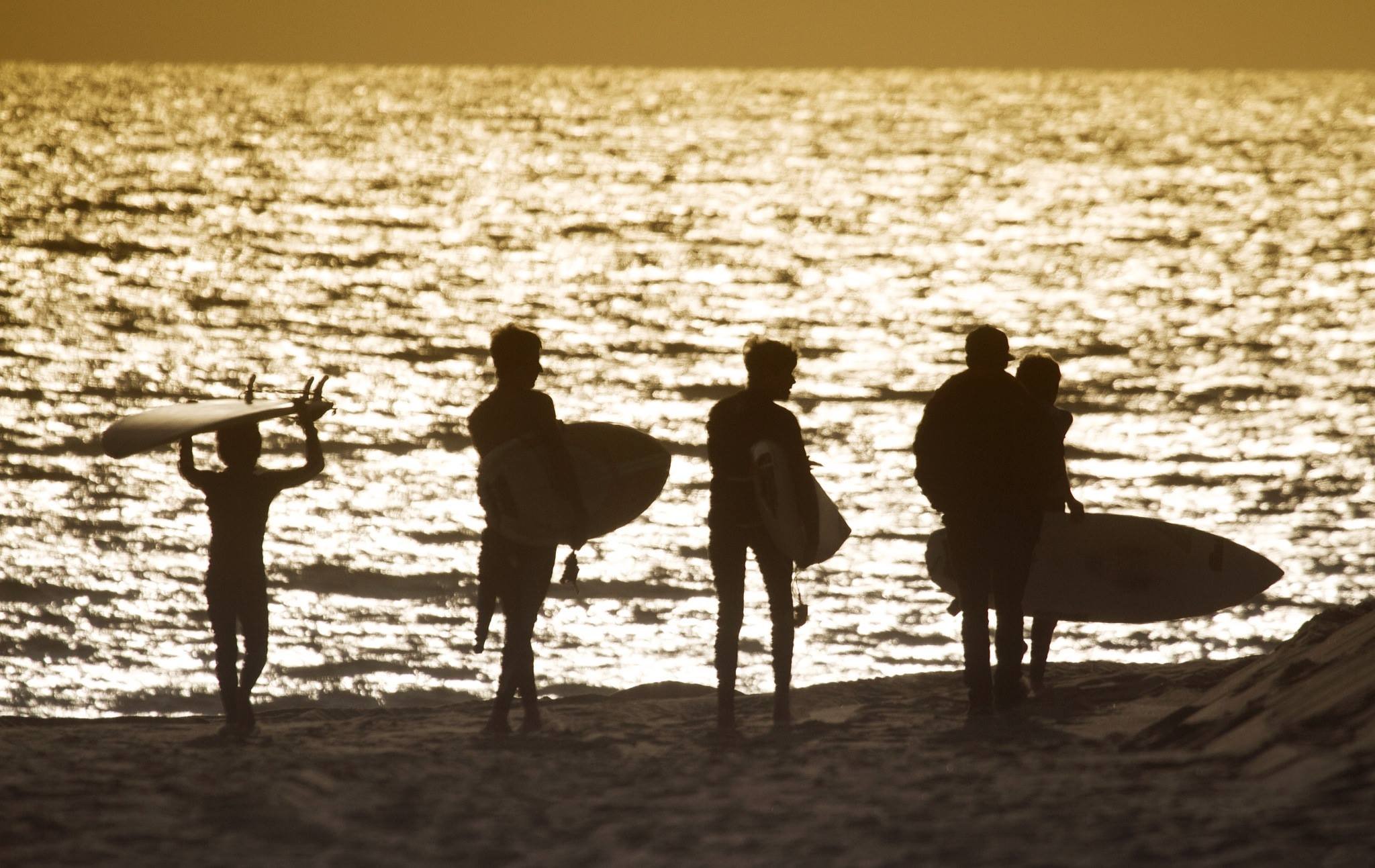 Cuatro personas de espaldas en la playa con tablas de surf, en Silueta, con mar brillante de fondo al atardecer.