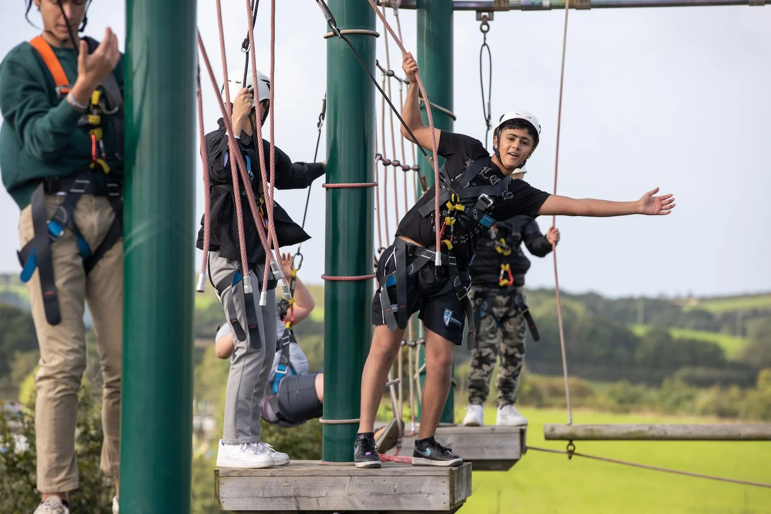 Niños con arneses en un parque de aventuras, algunos sobre plataformas de madera y una de ellas en un puente colgante con cuerda, en un entorno natural con hierba y colinas verdes.