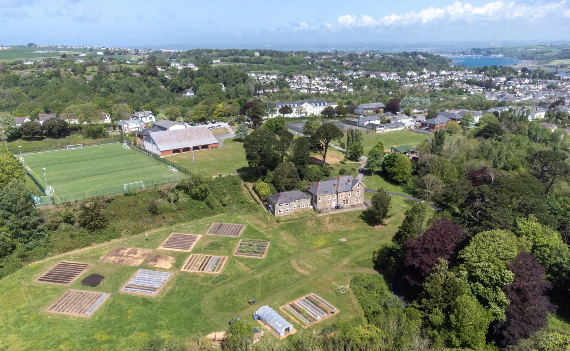 Vista aérea de un área verde con campos de cultivo, edificios residenciales y una zona de deportes con campos de fútbol, rodeado de árboles y colinas en el fondo.