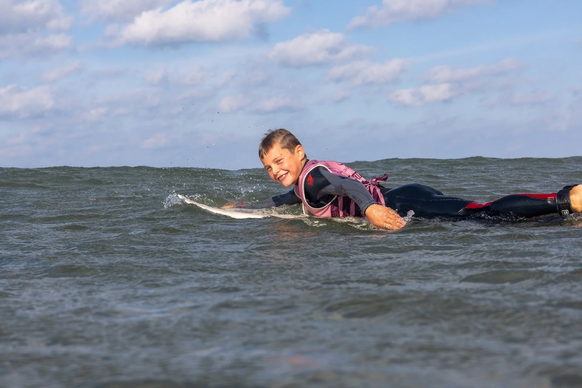 Niño disfrutando de surf en el océano con expresión feliz, usando traje de neopreno y una mochila en la espalda, bajo un cielo parcialmente nublado.