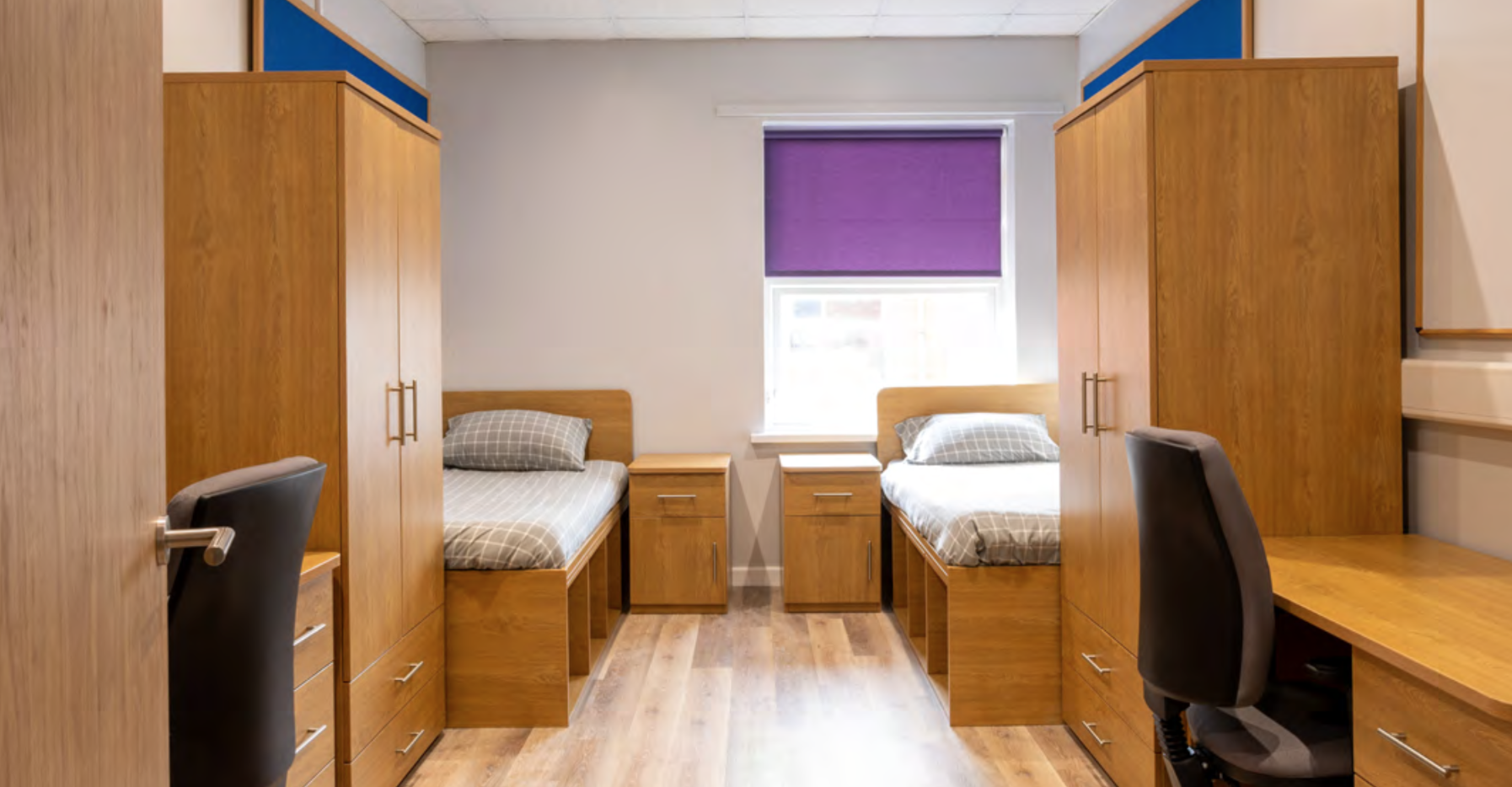 Dormitorio con dos camas individuales, armarios de madera, mesitas de noche, escritorios con sillas, y una ventana con persiana morada