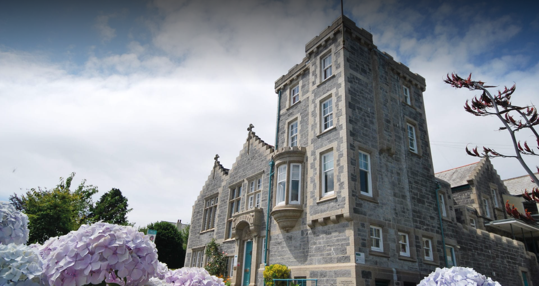 Edificio antiguo de piedra con ventanas grandes y torre en medio, rodeado de flores de hortensias en primer plano y árboles verdes bajo un cielo parcialmente nublado.