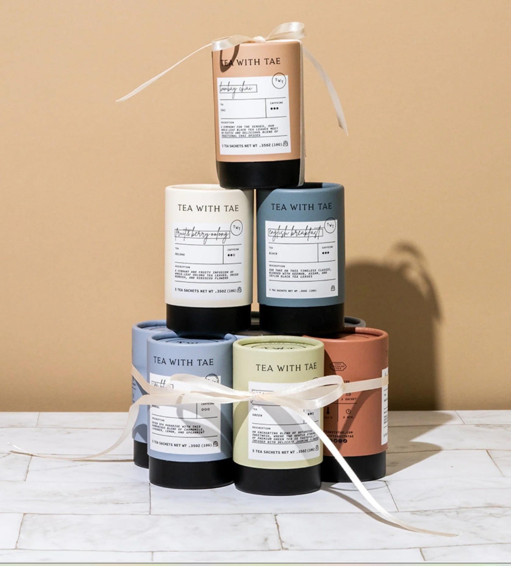 Tea with Tae - Bedtime Bliss: Stack of seven colorful, cylindrical tea packages tied with white ribbon, resting on a marble surface.