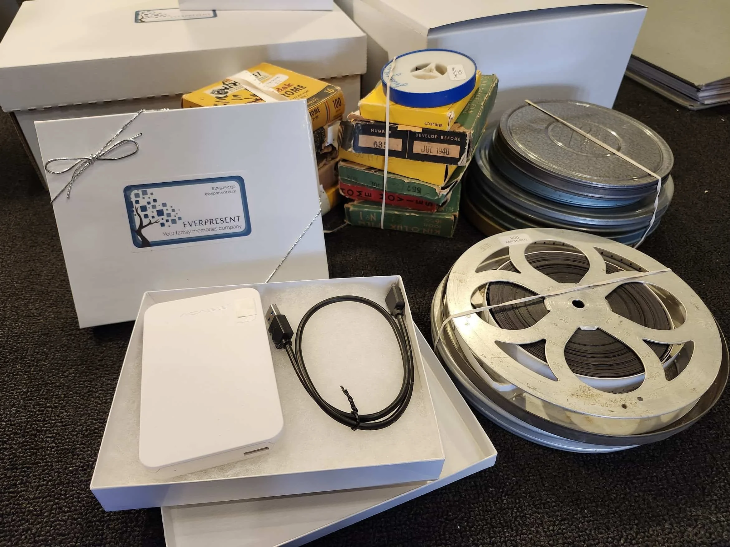 Media transfer kit in a white box with the EverPresent logo, an external hard drive and cable, surrounded by stacks of vintage film reels and tapes.