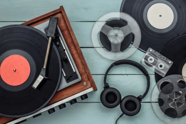 Overhead view of a vintage wood turntable with a black vinyl record, headphones, and a reel-to-reel tape on a pale blue wooden surface.