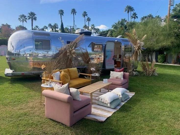 Outdoor setup with pastel-colored sofas, wooden table, pillows, and dried plants in front of a shiny silver trailer on a grassy area, with palm trees and blue sky in the background.