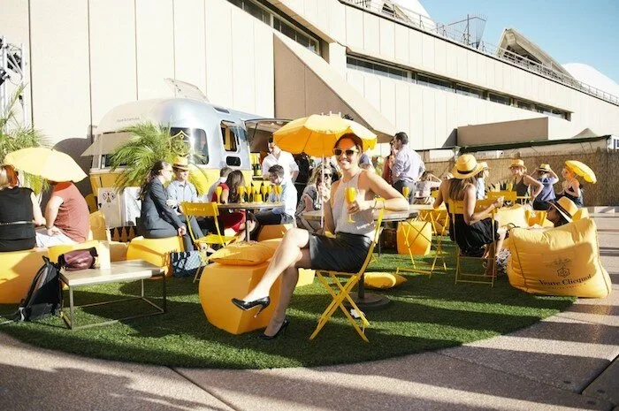 People gathered outdoors on yellow chairs and bean bags, with yellow umbrellas, near a silver trailer, enjoying drinks on a sunny day.
