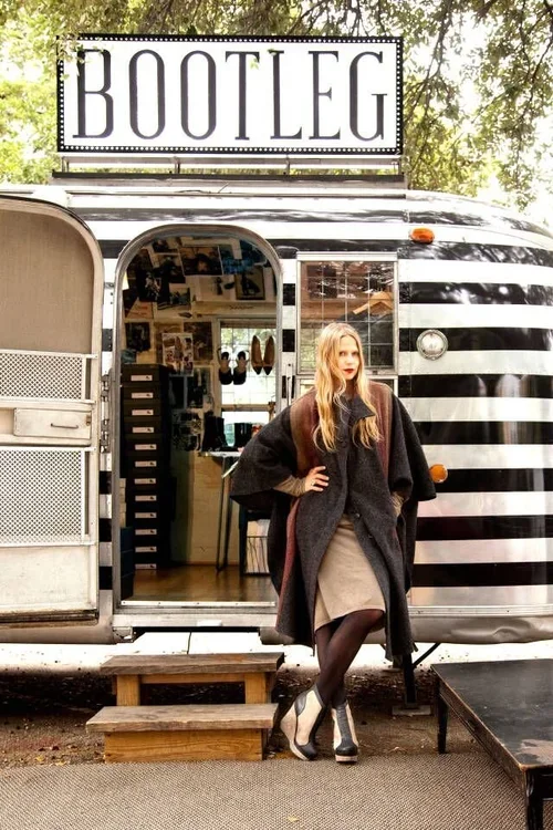 A woman standing outside a black and white striped mobile boutique trailer with a sign that says 'BOOTLEG' on top, surrounded by trees.