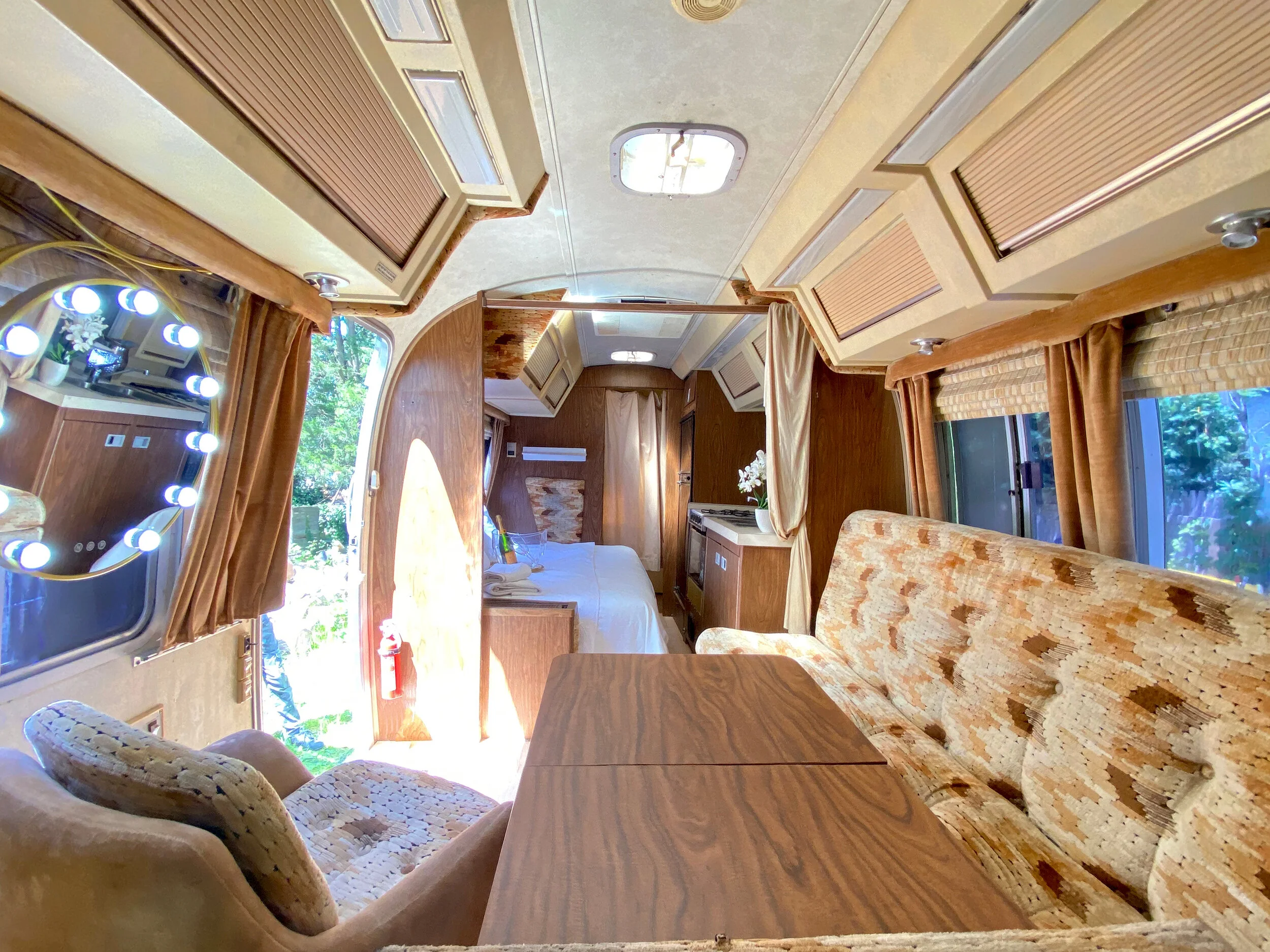 Interior of a vintage camper trailer with wood paneling, a sofa with a patterned upholstery, a small table, a bed with white linens, and a window with brown curtains.