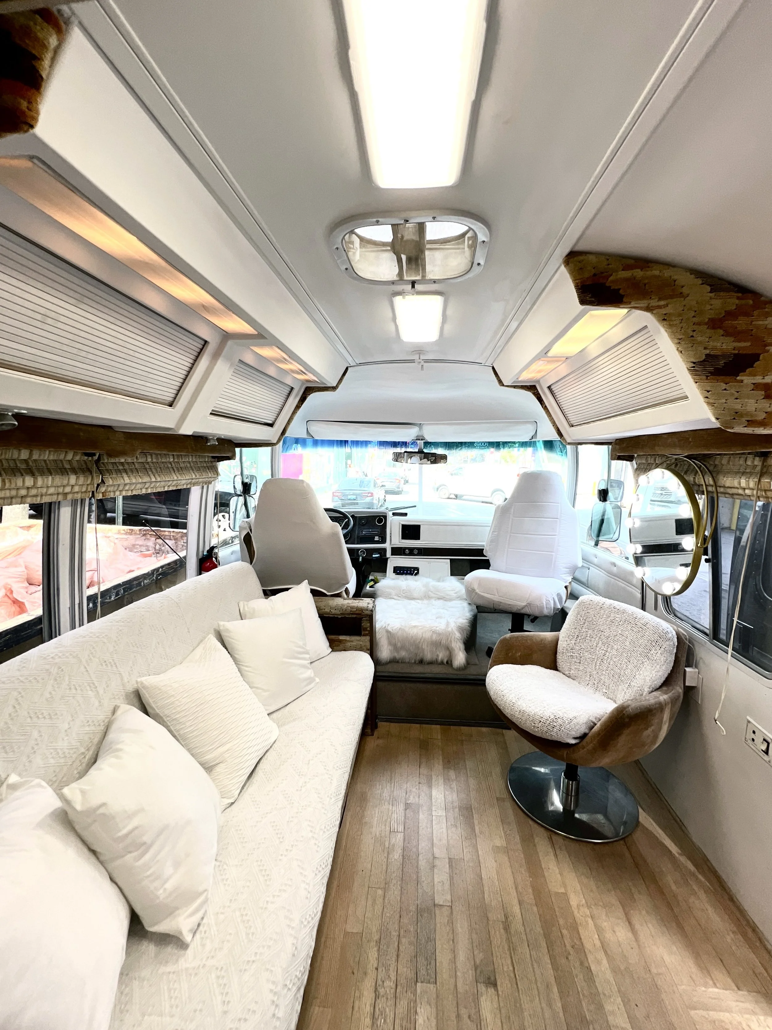 Interior of a converted bus with a comfortable seating area, including a cream-colored sofa with white pillows, a brown swivel chair, and two white captain seats at the front. The ceiling has built-in lighting and a skylight, and storage cabinets are