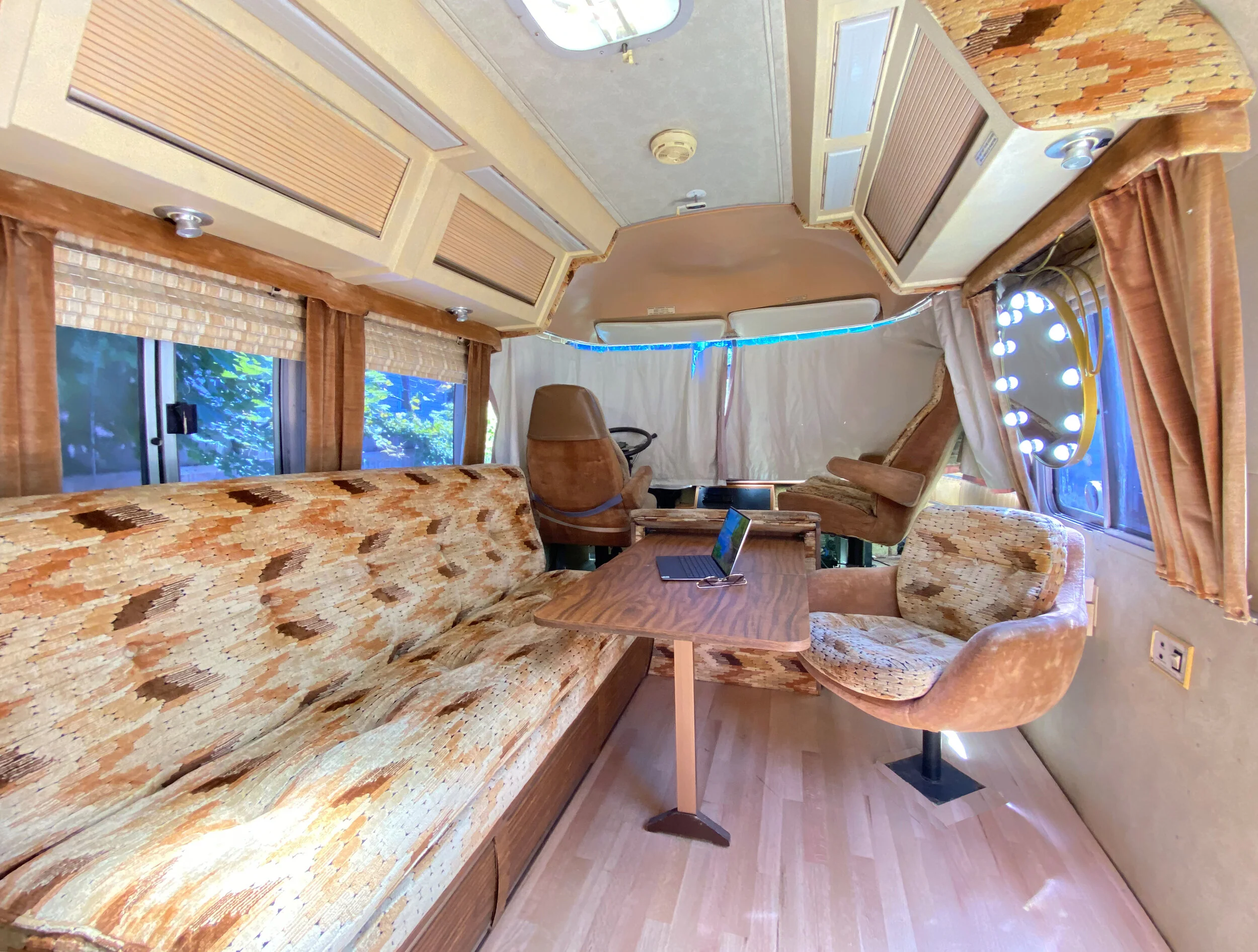 Interior of a vintage RV with patterned upholstery on a sofa and chairs, wooden table, windows with beige curtains, and ceiling storage compartments.